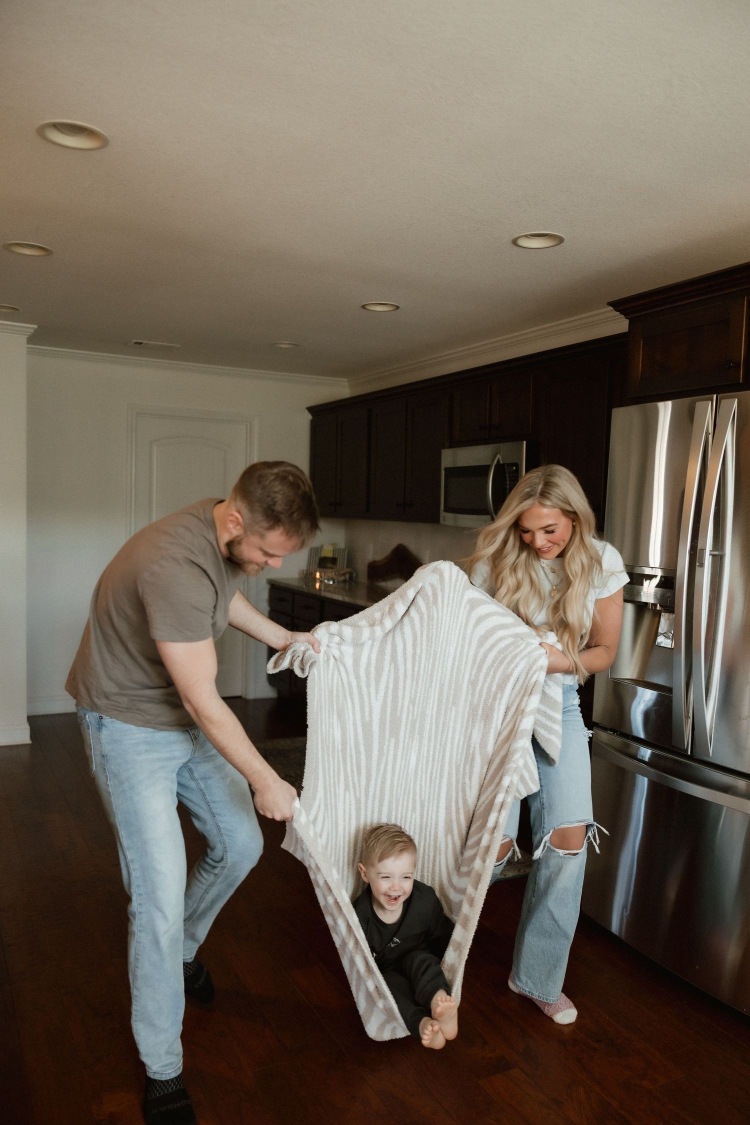 Parents playfully wrapping laughing toddler in striped blanket in modern kitchen with dark cabinets.