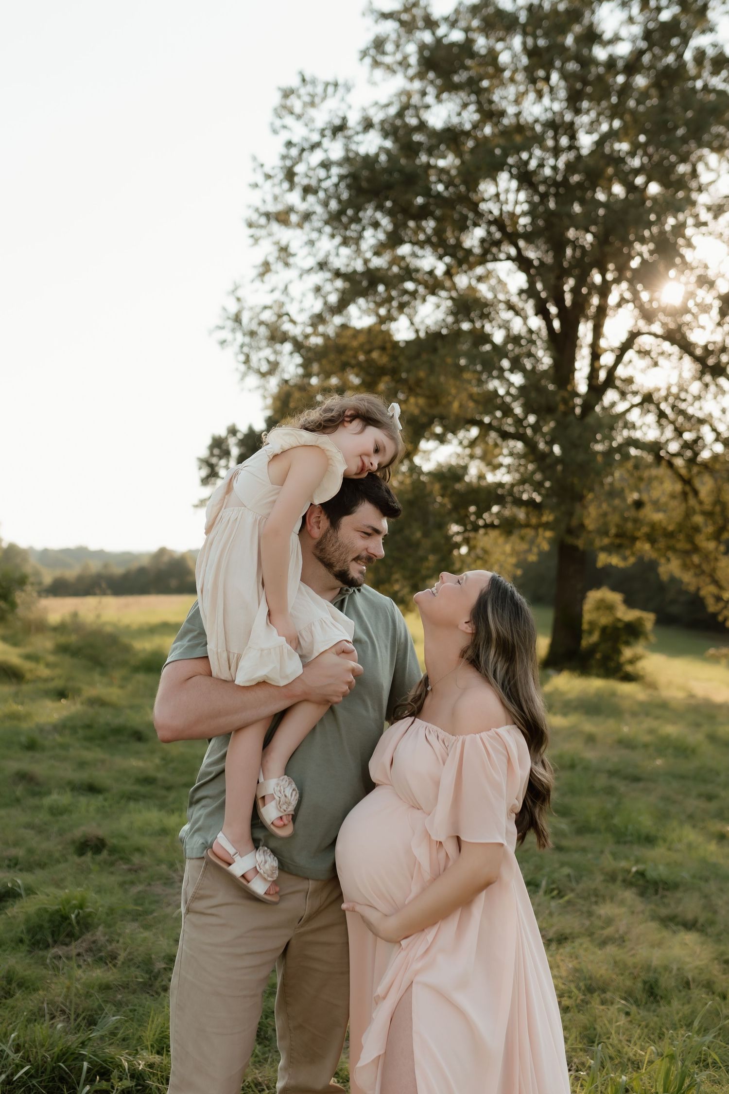 Expecting couple holding young daughter at golden hour in a lush green field with sunlit tree backdrop.
