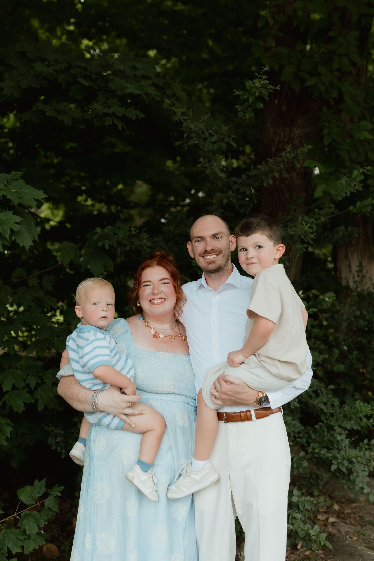 Happy family of four with two young boys posing outdoors in coordinating light blue and neutral outfits.