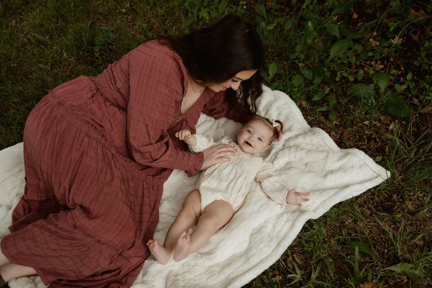 Mother in rust dress lying on blanket with baby in garden.
