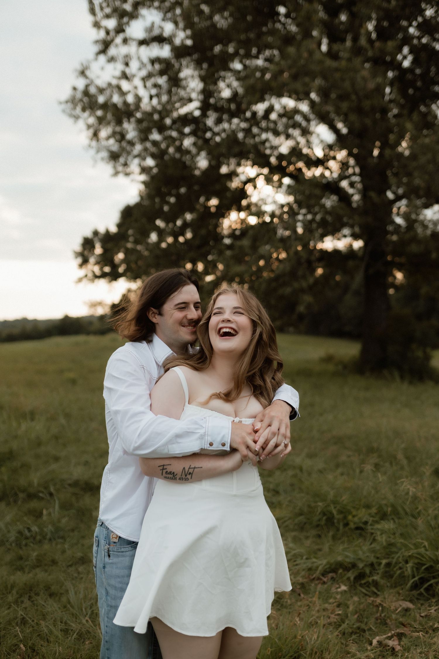 Joyful couple embracing in green meadow at sunset, woman in white dress laughing while man hugs her from behind.