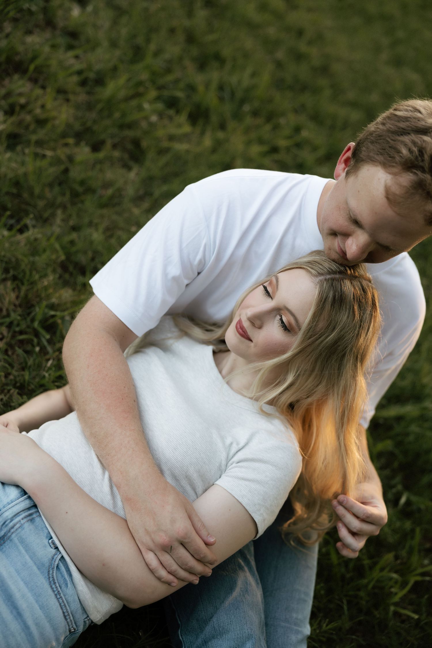 A man tenderly kisses a woman's head while embracing her as she leans back against him on the grass.