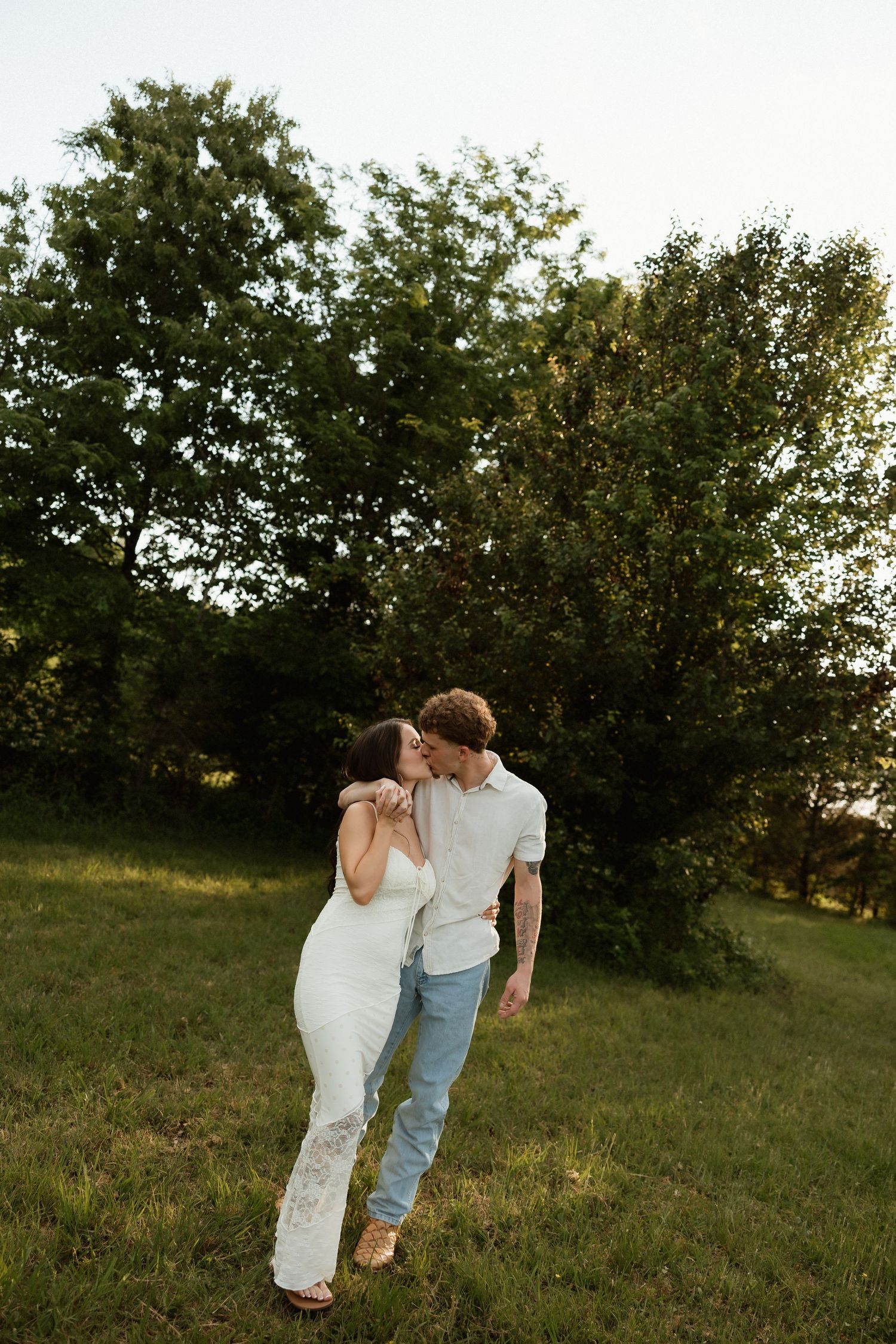 A couple shares a romantic kiss on a sunny green lawn with trees in the background.