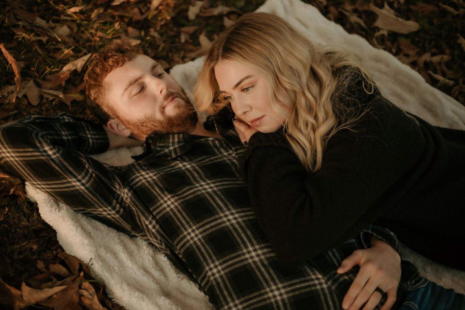 Couple lying together on plaid blanket surrounded by autumn leaves in cozy outdoor engagement session.