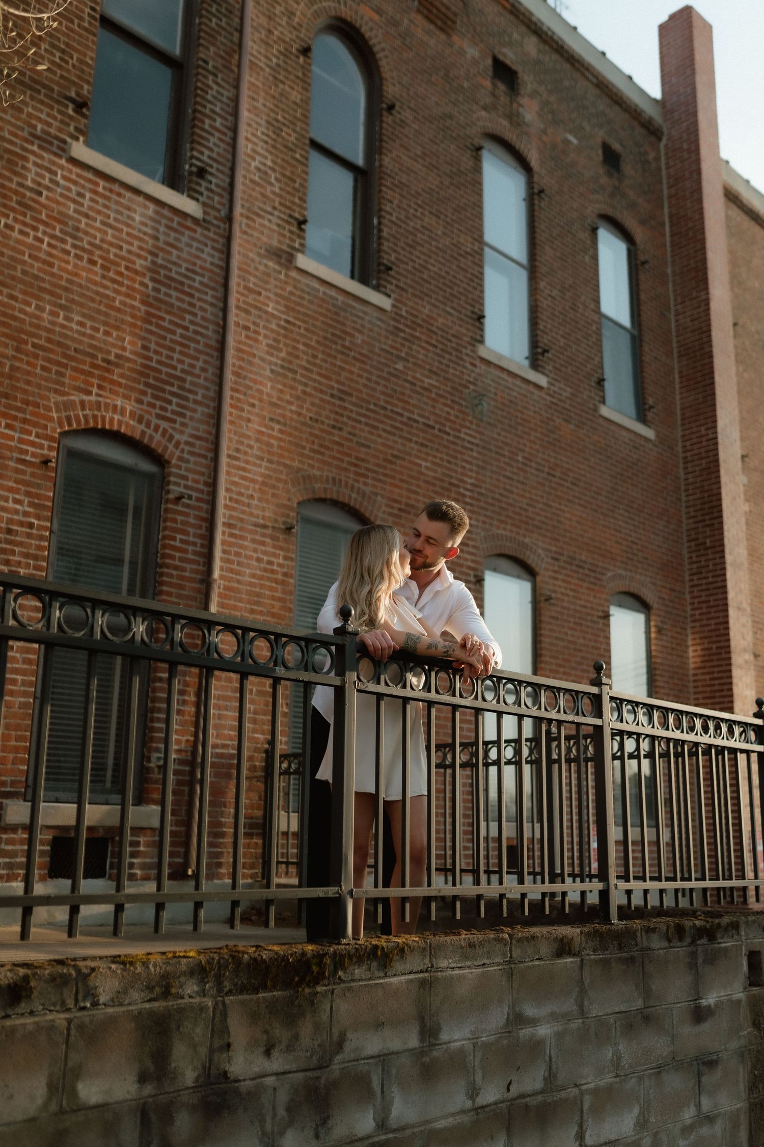 Romantic couple embracing on decorative iron fence balcony against classic red brick building facade.