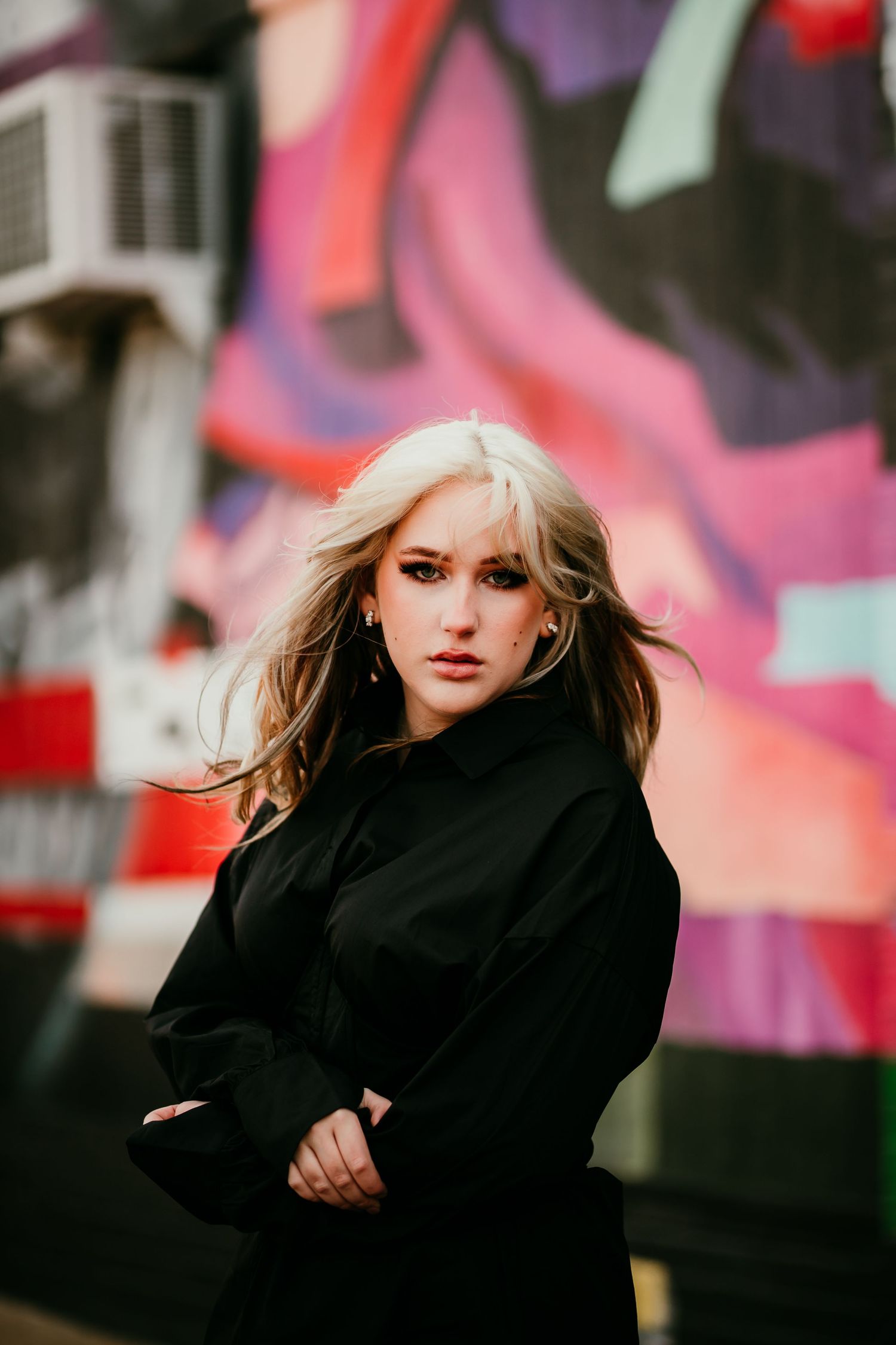 Blonde woman in black outfit posing before colorful graffiti wall.