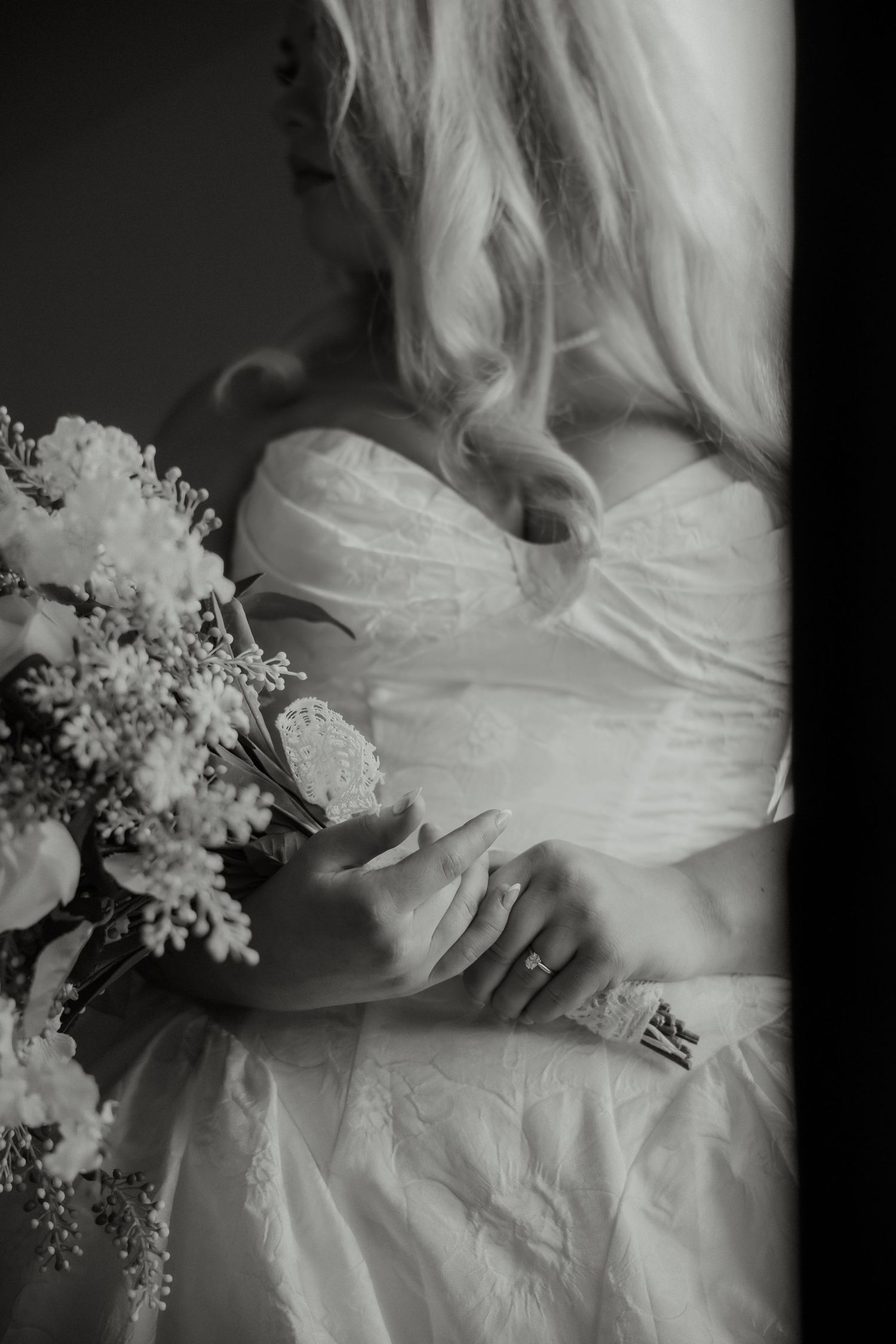 Black and white photo of blonde bride in lace gown holding white floral bouquet indoors.
