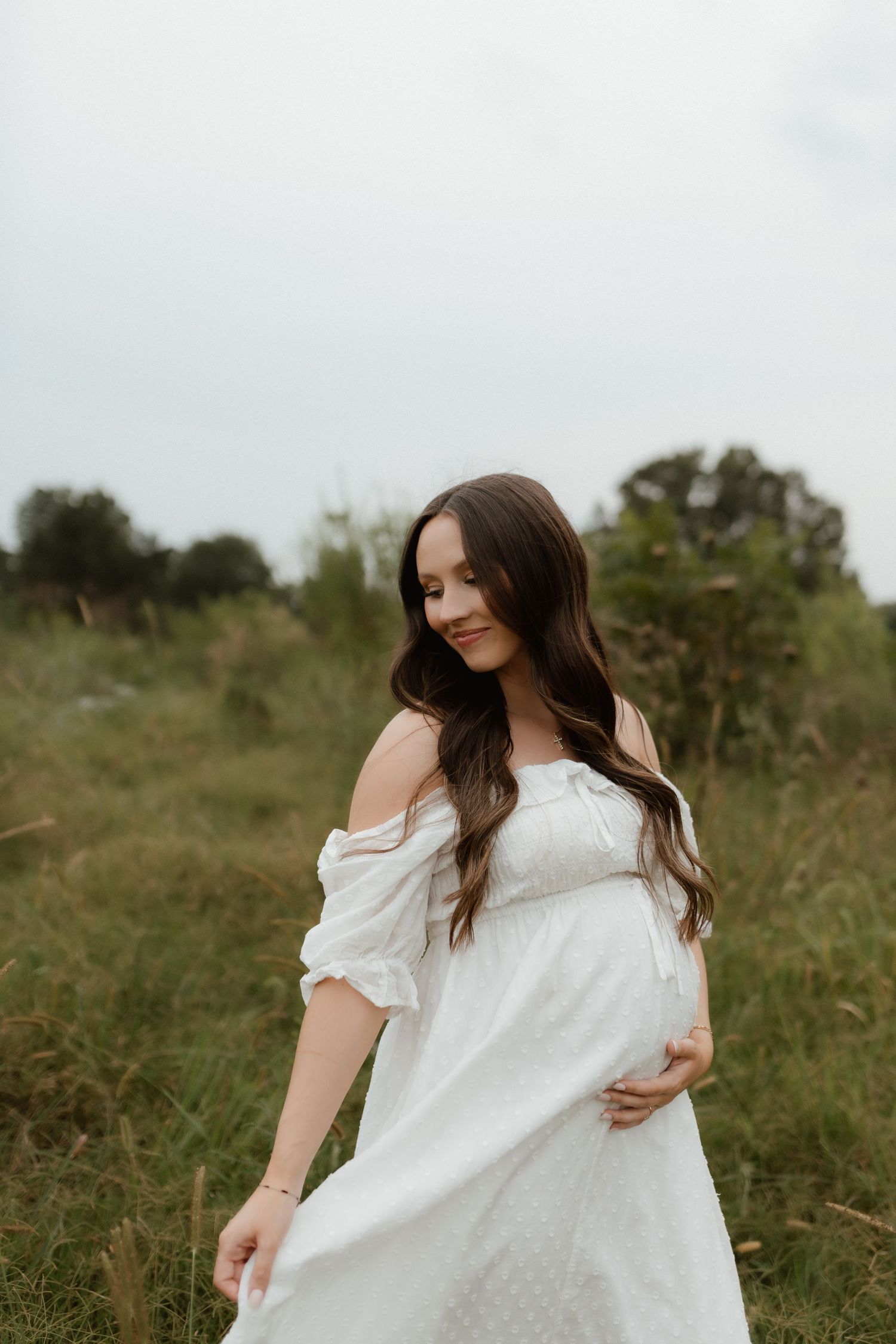 Pregnant woman in white off-shoulder dress cradles baby bump while standing in green field.