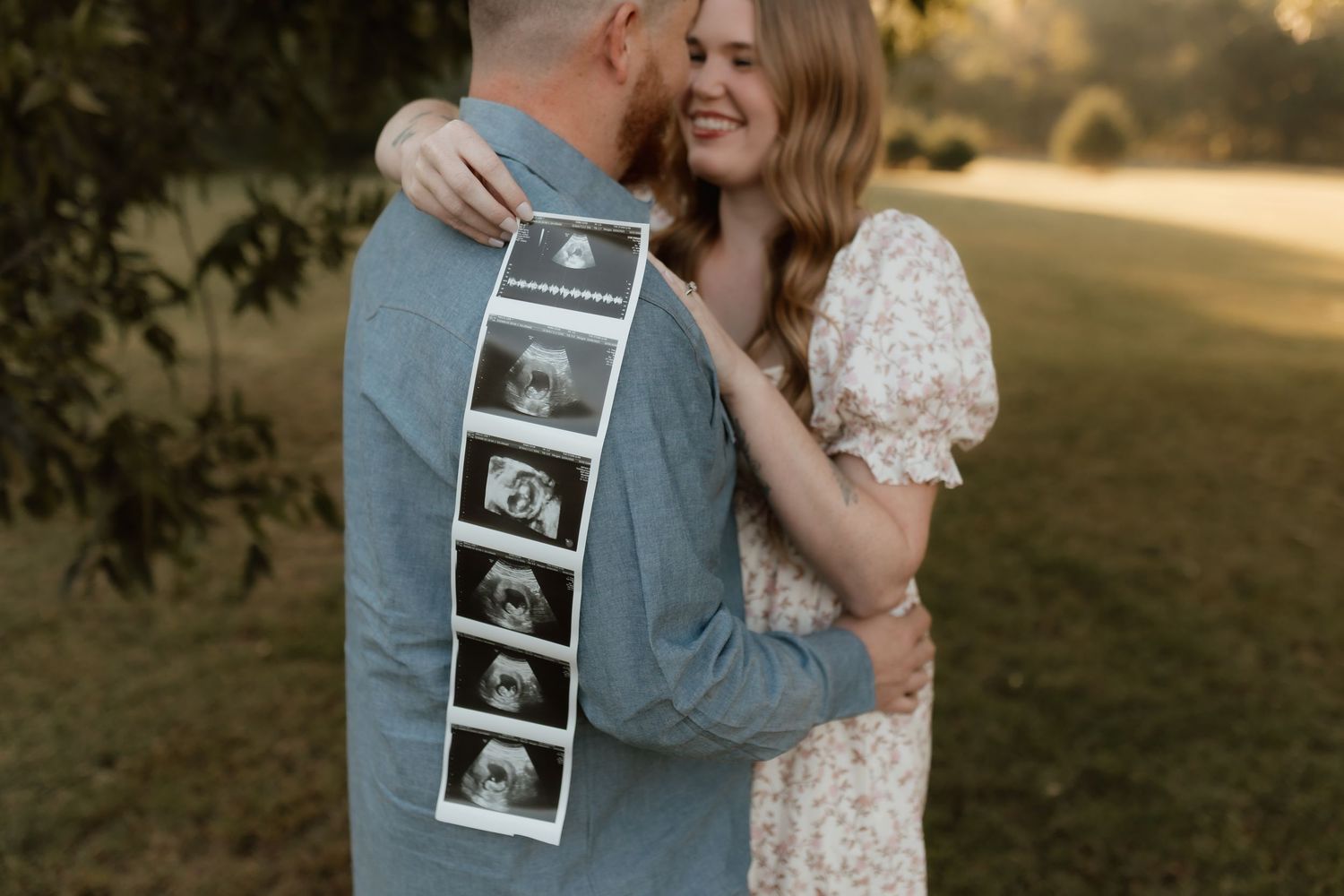 Couple embraces outdoors holding ultrasound photos, announcing pregnancy in a garden setting.