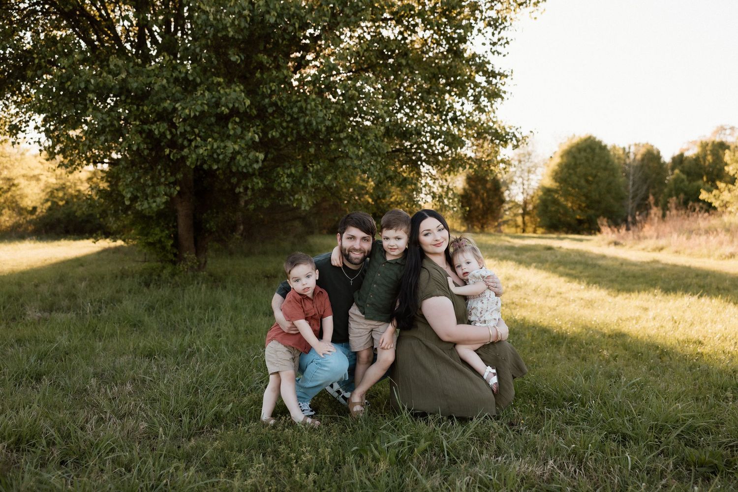Happy family of five posing together on green grass with a large tree in background at sunset.