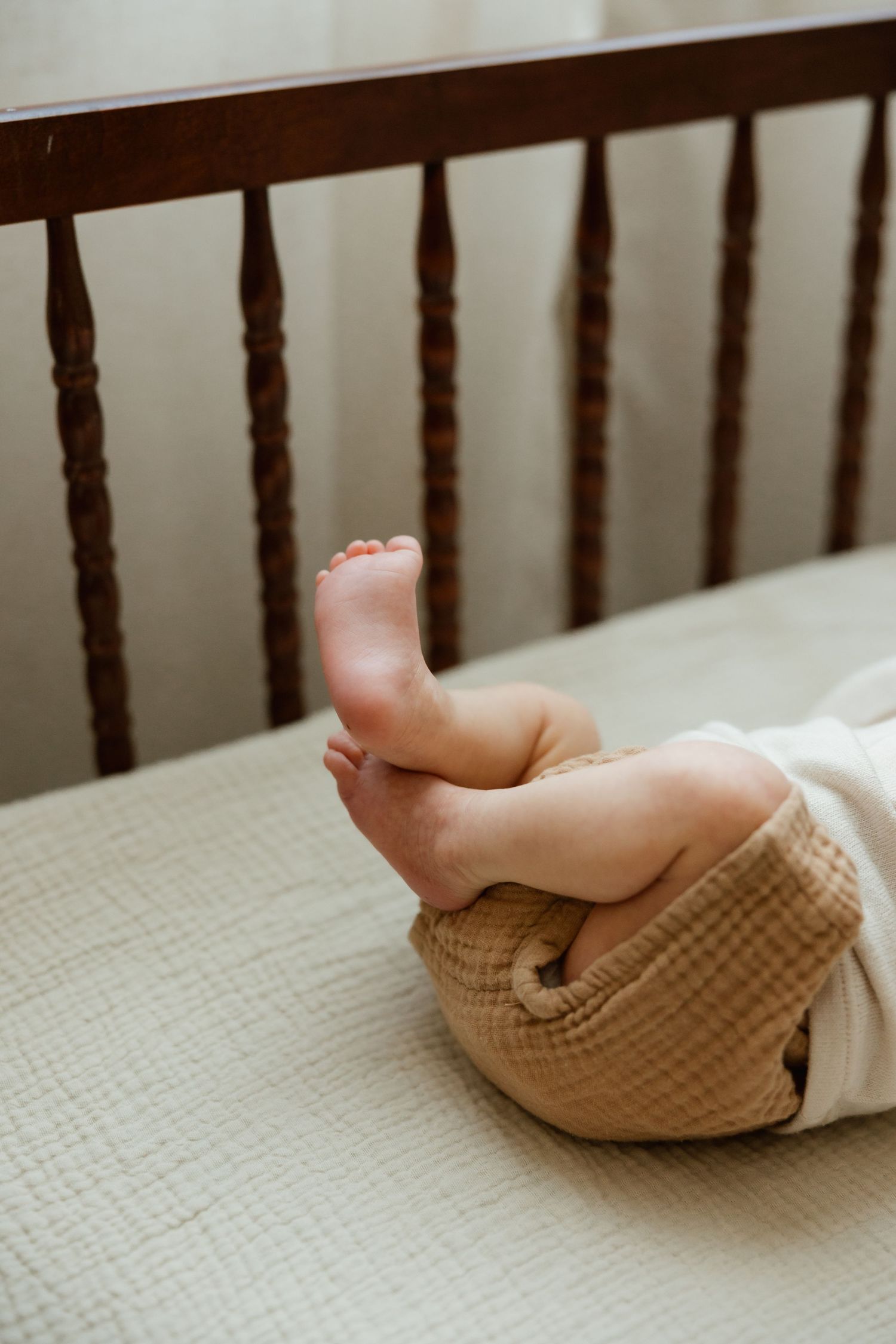 Close-up of newborn baby feet and legs in tan bloomers resting in a wooden crib.