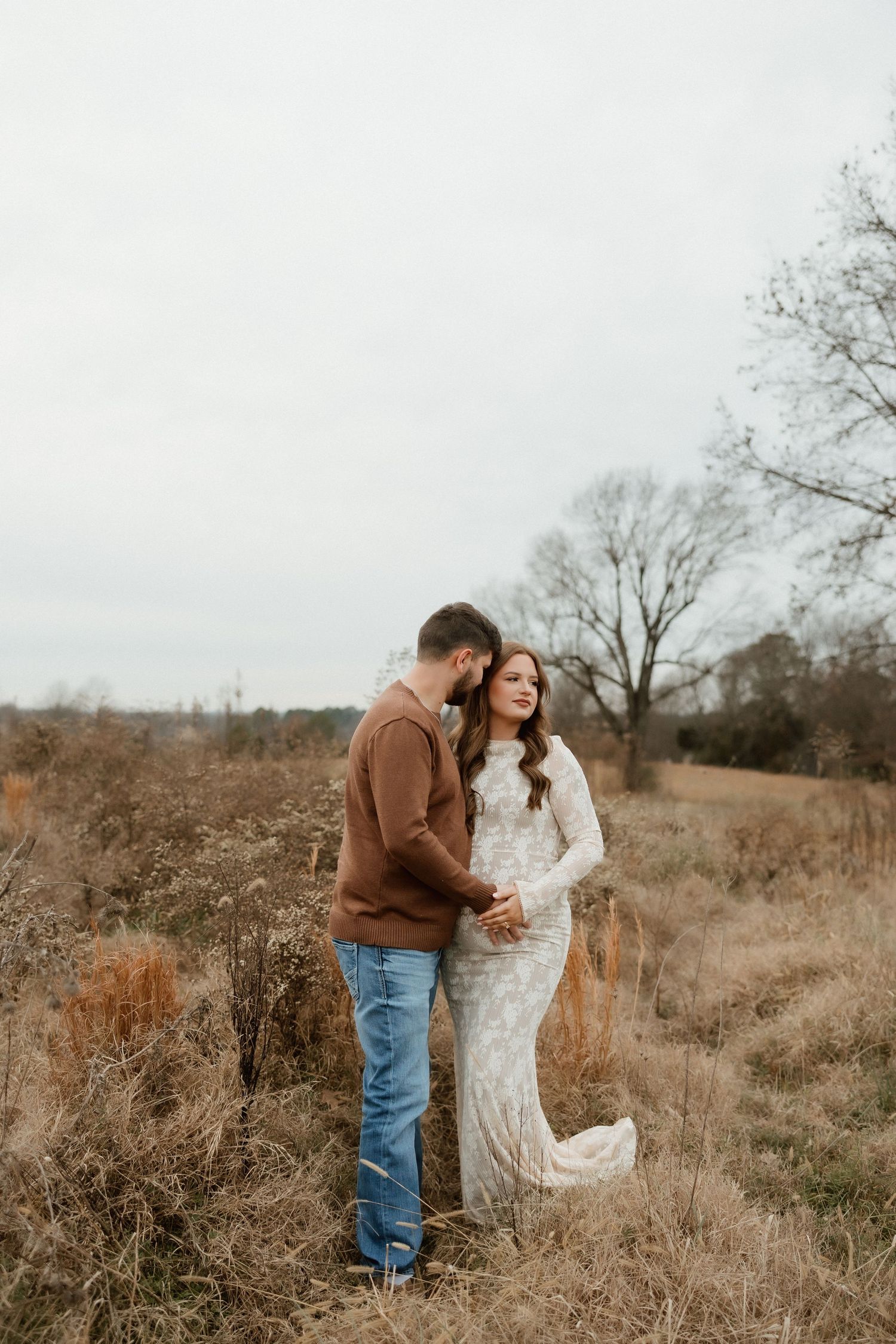 Couple embracing in a golden field during maternity photoshoot, woman in white lace gown, man in brown sweater and jeans.