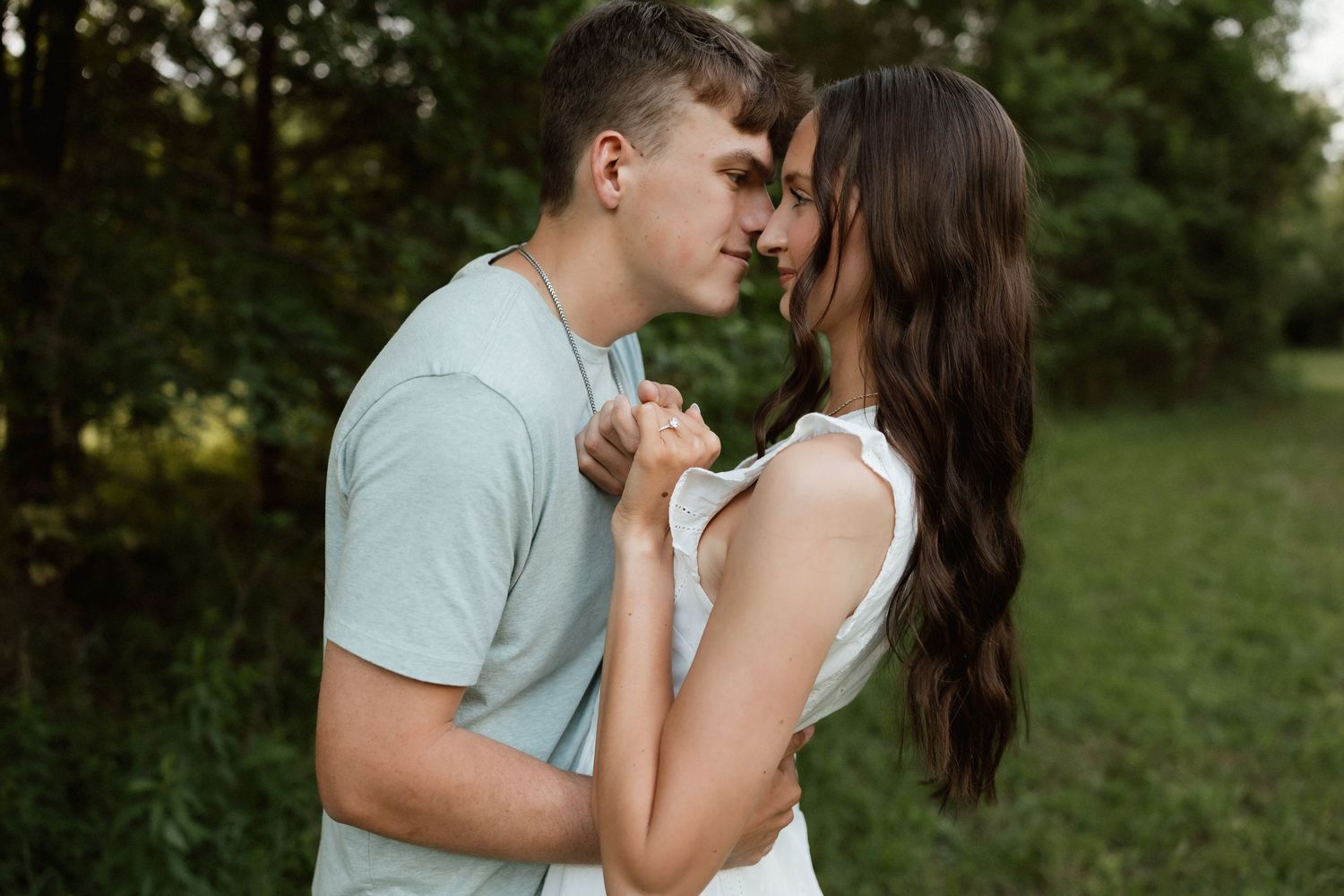 A young couple with foreheads touching, holding hands tenderly in a lush green outdoor setting.