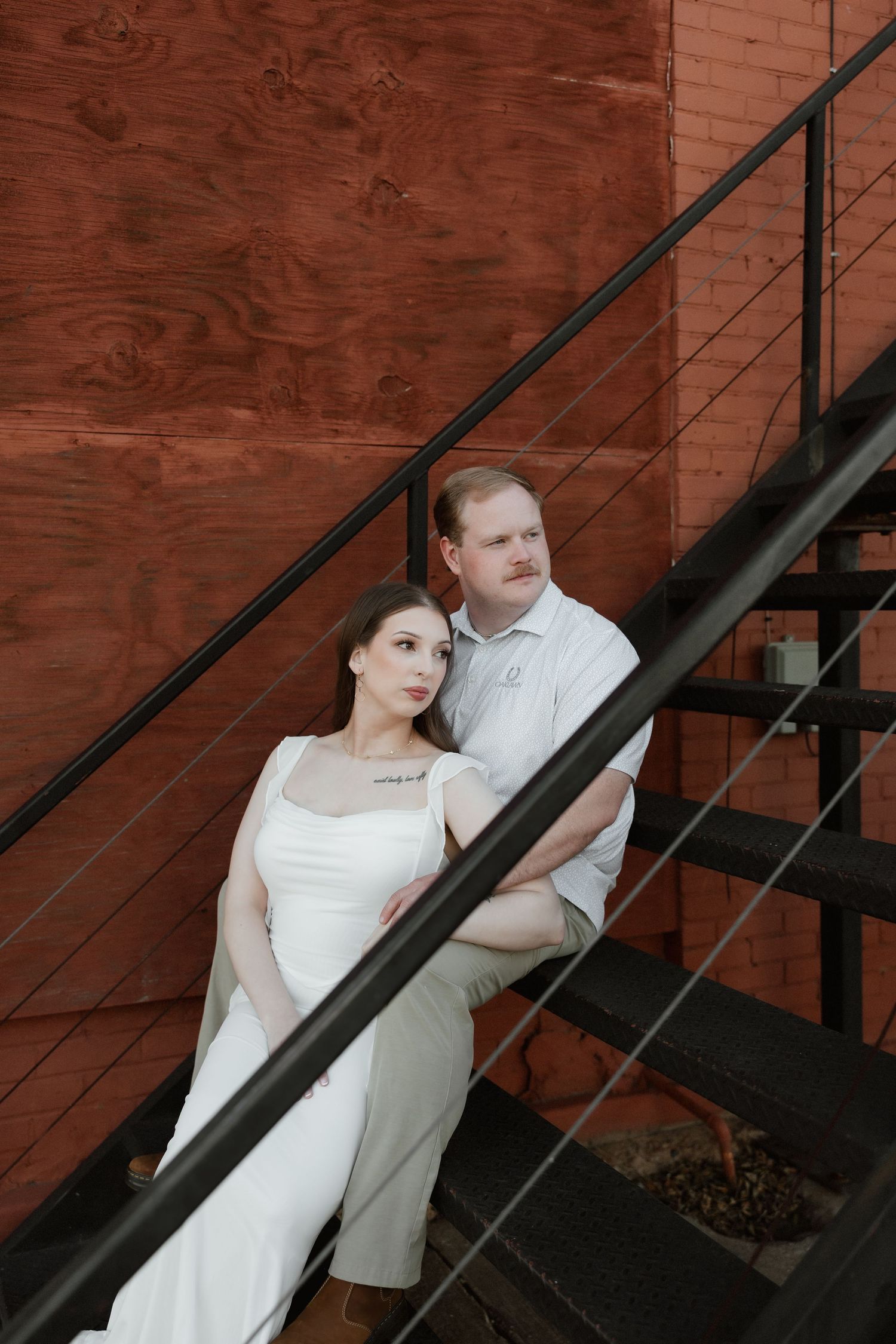 An elegant couple posing on an outdoor staircase against a rustic red wood panel wall.