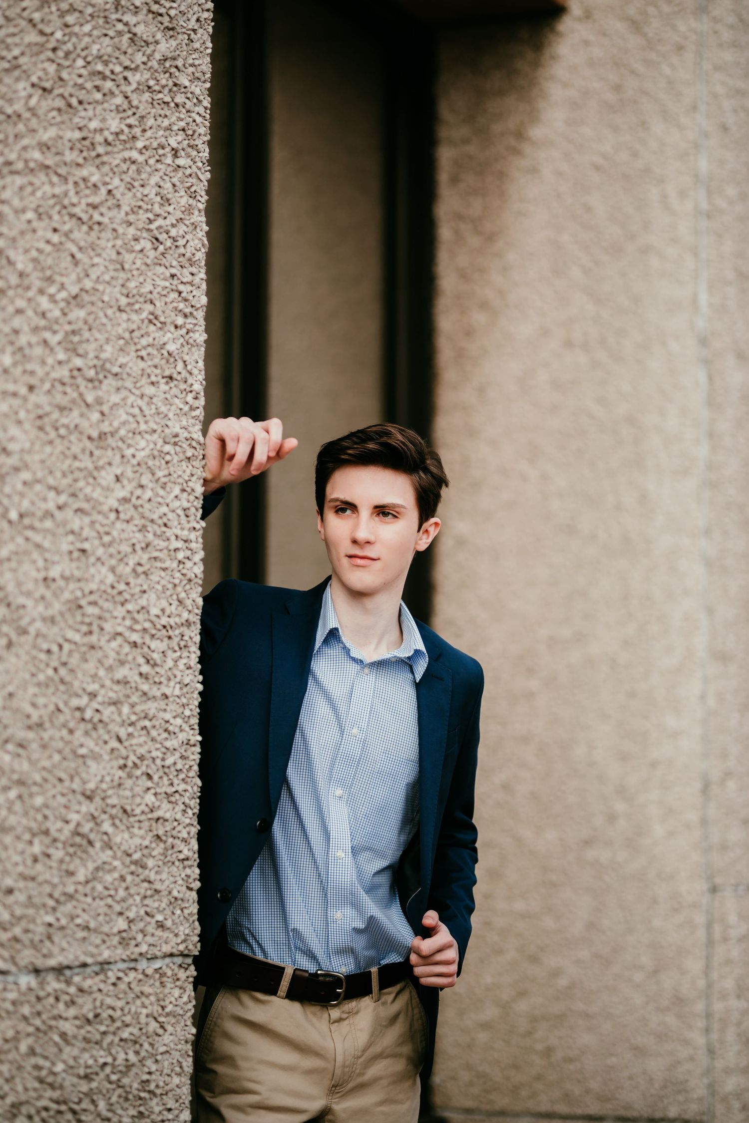 Young man in navy blazer and khakis leans against textured stone wall outdoors.