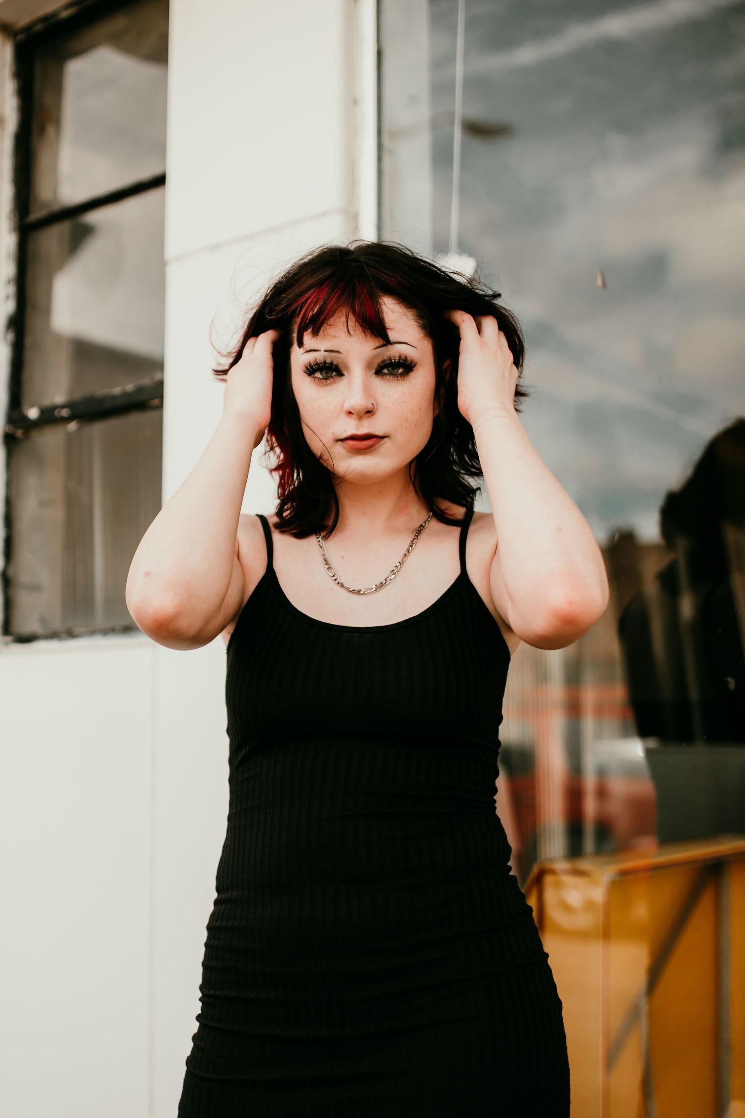 Young woman in black dress with dramatic makeup holding her dark hair beside a storefront window.