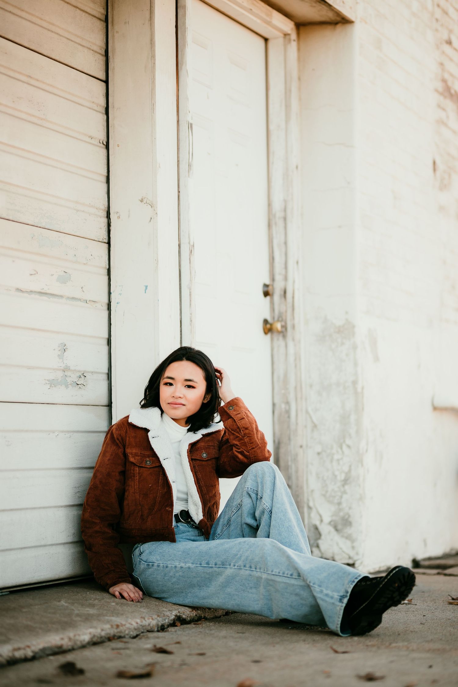 Young Asian woman in brown sherpa jacket sitting against white wooden doors on concrete ground.