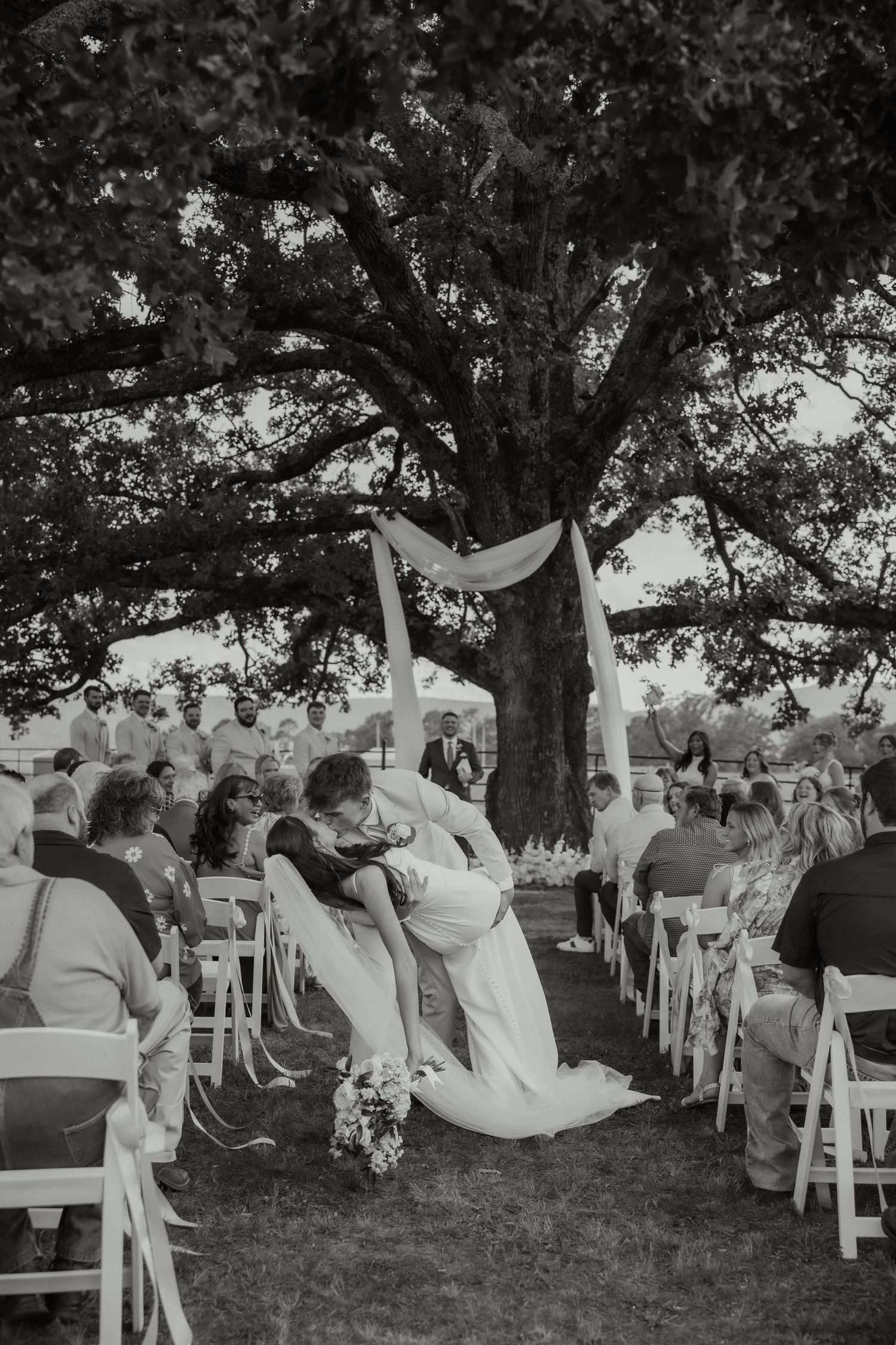 Black and white photo of bride and groom kissing at outdoor ceremony under large oak tree.