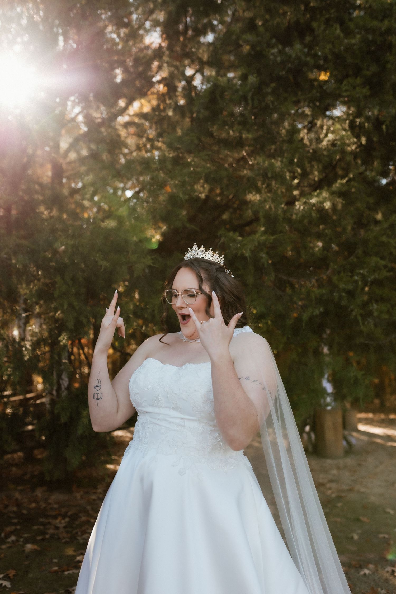 Excited bride in white wedding gown and crown makes rock on gesture outdoors in golden sunlight.