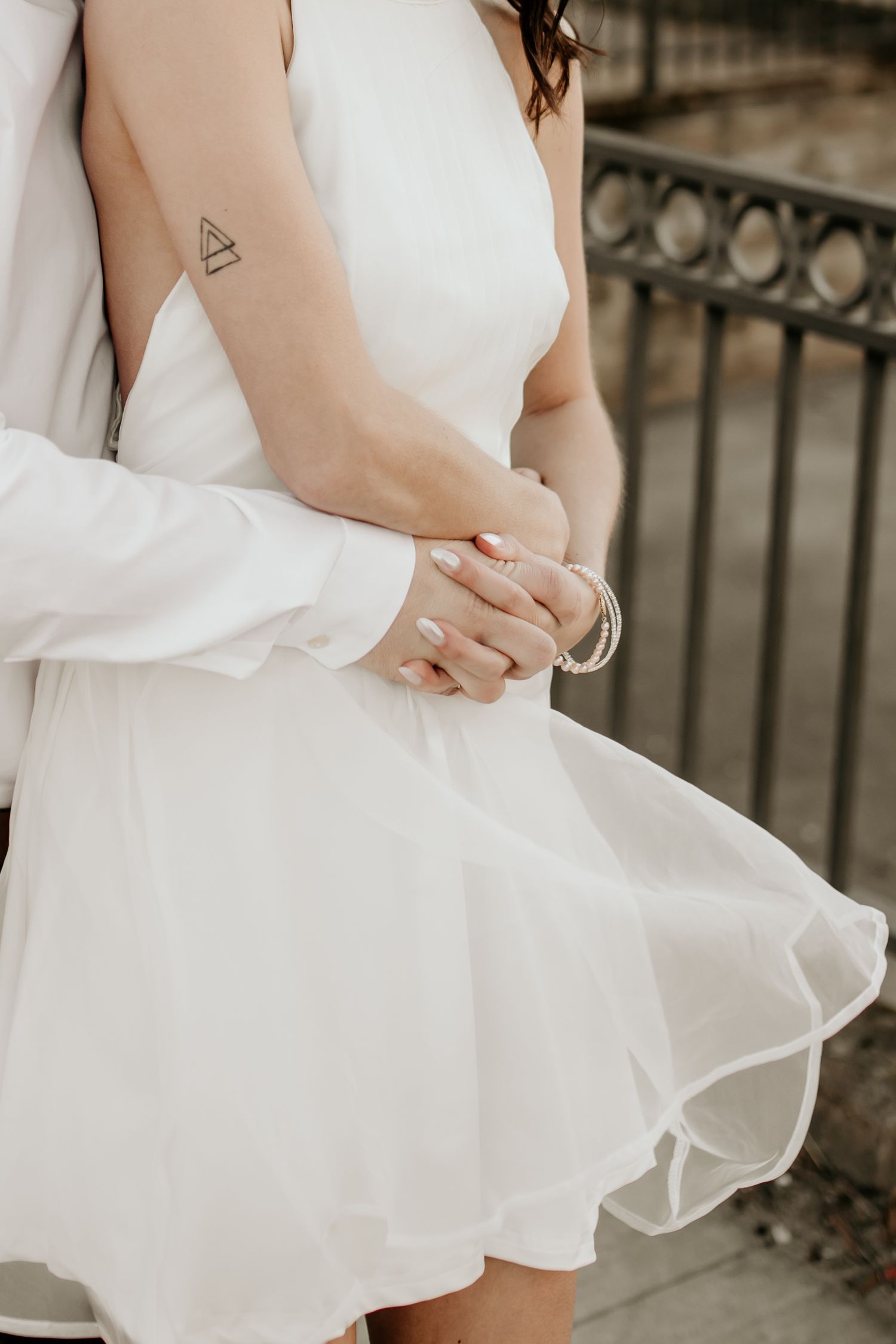Close-up of couple holding hands, woman in white dress with triangle tattoo and bracelet during engagement shoot.