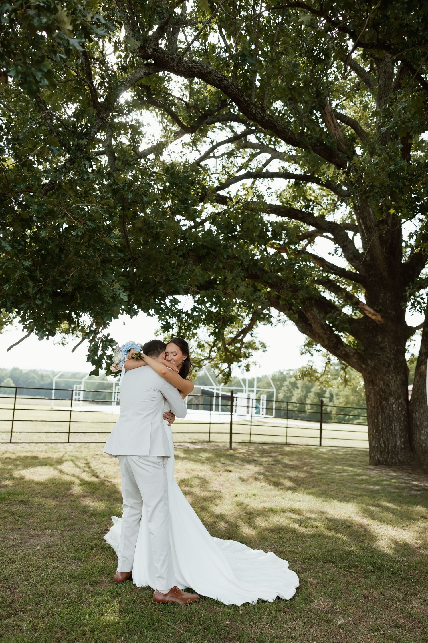 Bride and groom embracing under sprawling oak tree with farm fence visible in background.