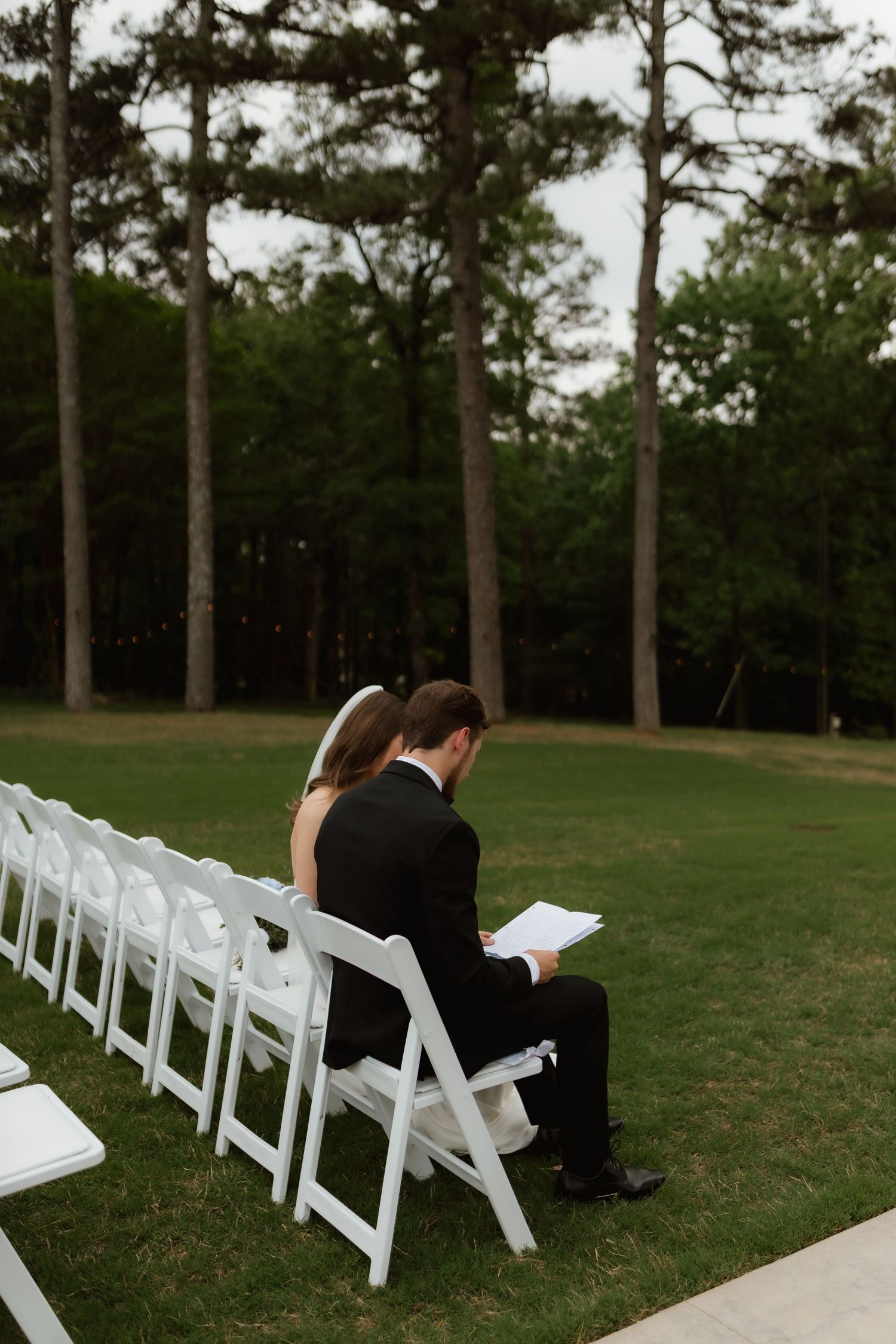 Bride and groom sit together reading wedding programs in white chairs at outdoor ceremony.