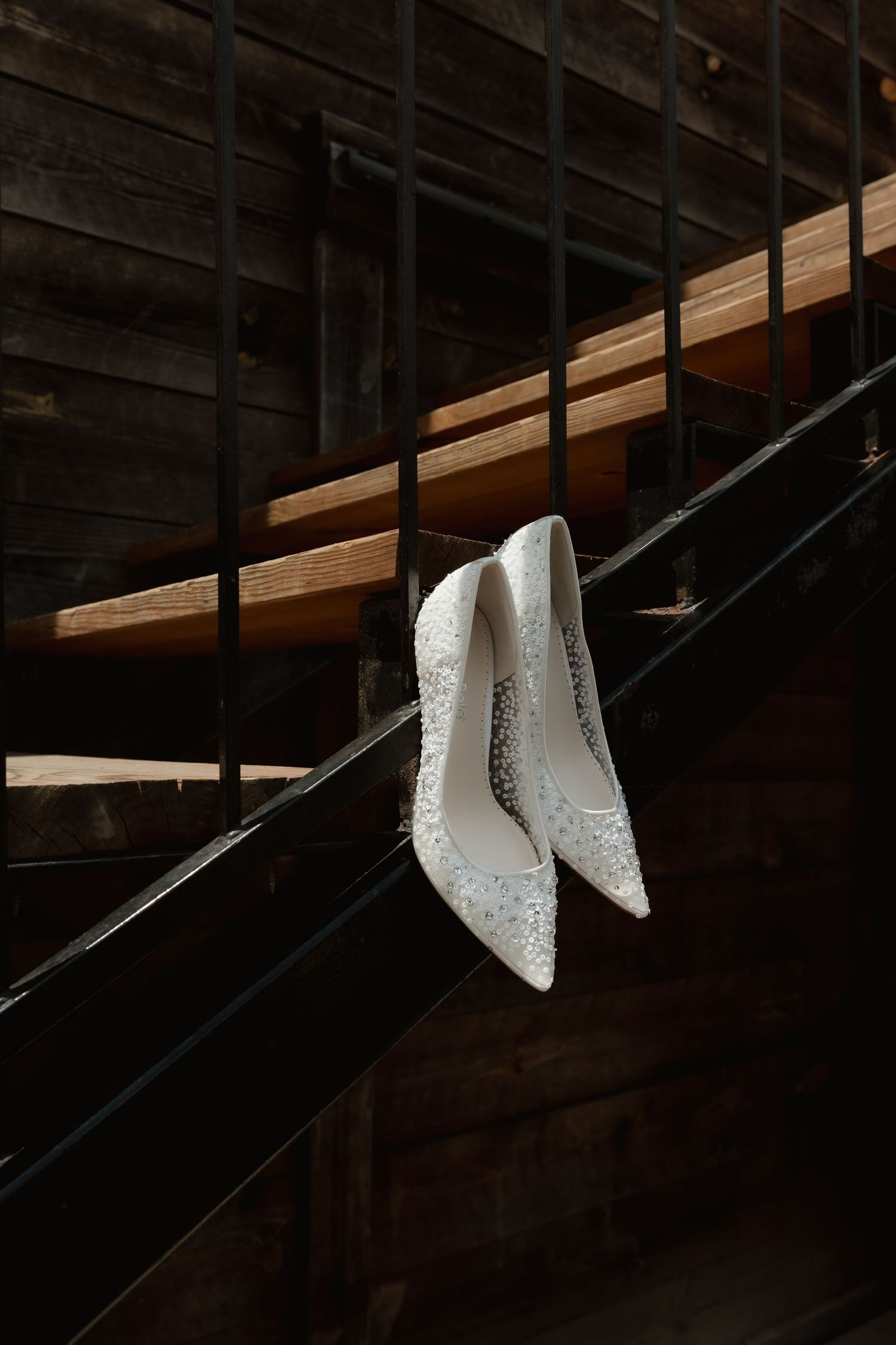 White crystal-embellished bridal heels hanging on dark staircase railing in rustic wooden venue.