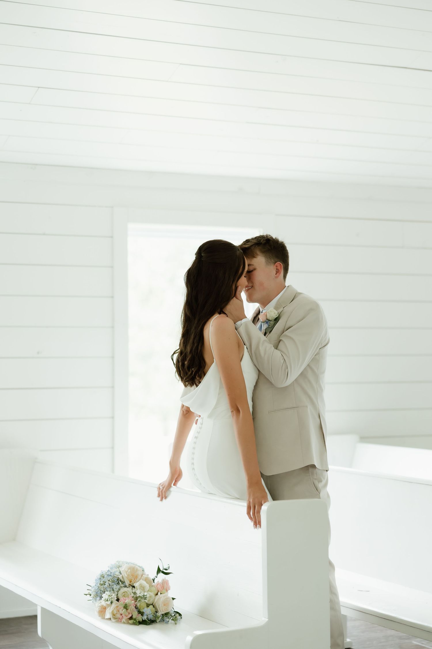 Bride and groom share a romantic kiss inside a bright white chapel with wooden pews.