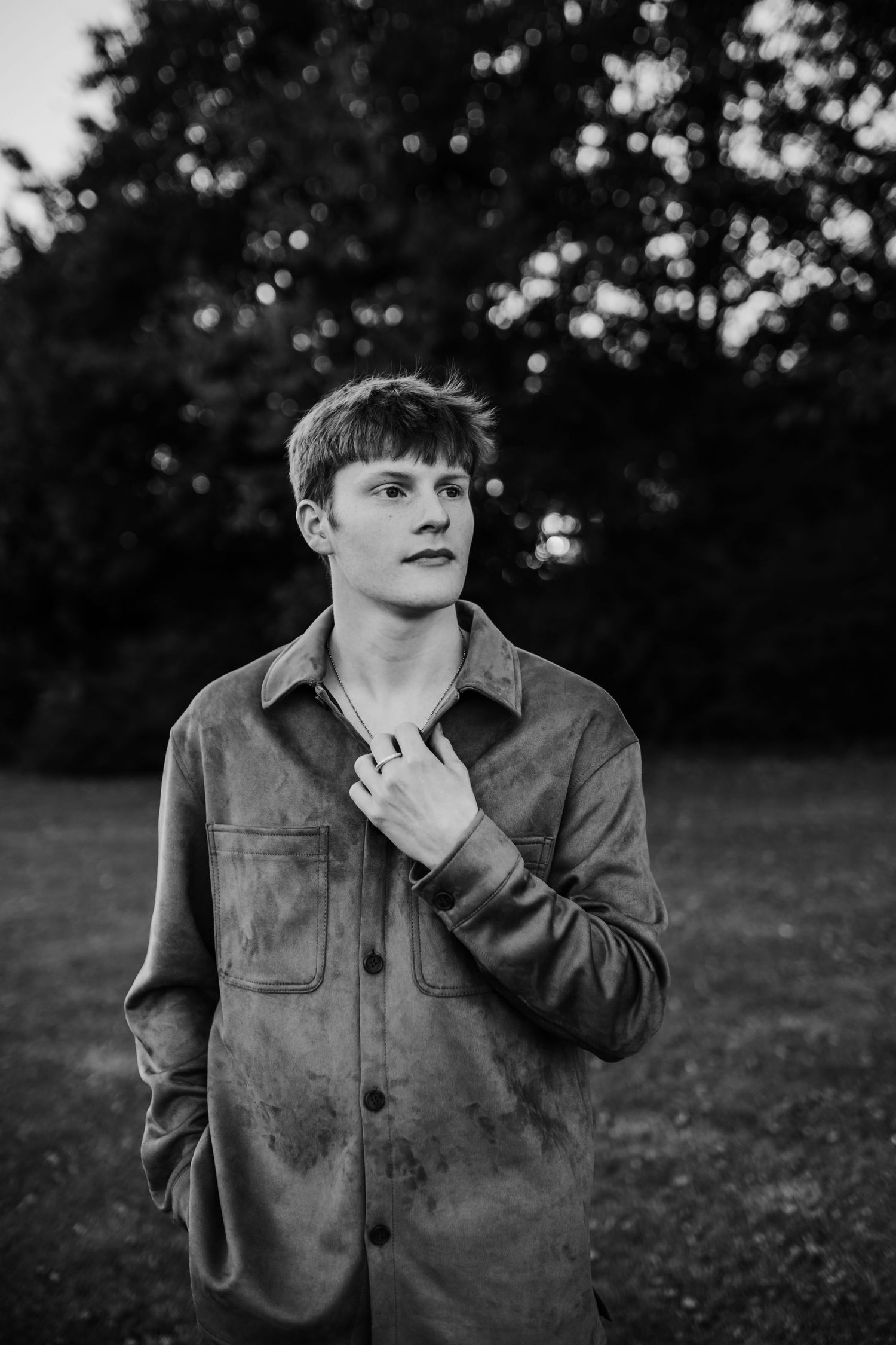 Black and white portrait of young man in button-up shirt standing outdoors among trees.