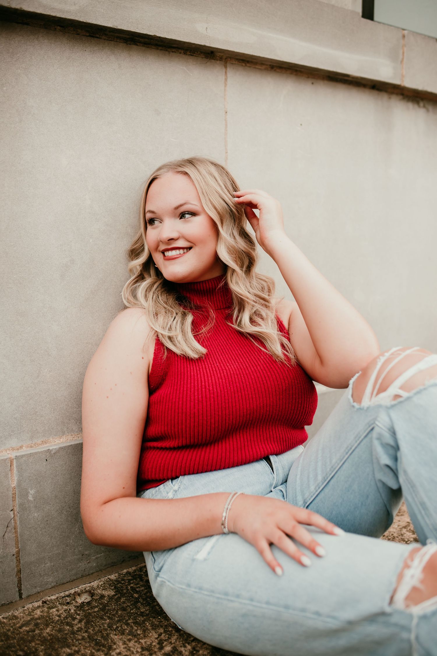 Smiling blonde woman in red sleeveless top and ripped jeans poses against concrete wall outdoors.