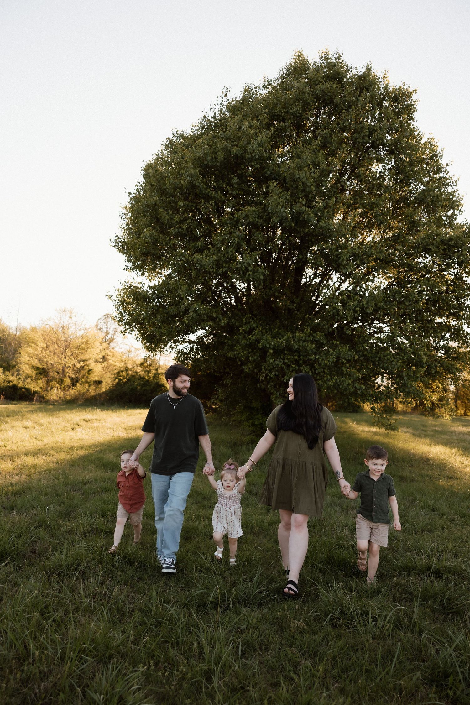 Family of five holding hands walking through a sunlit green field with a large tree backdrop.