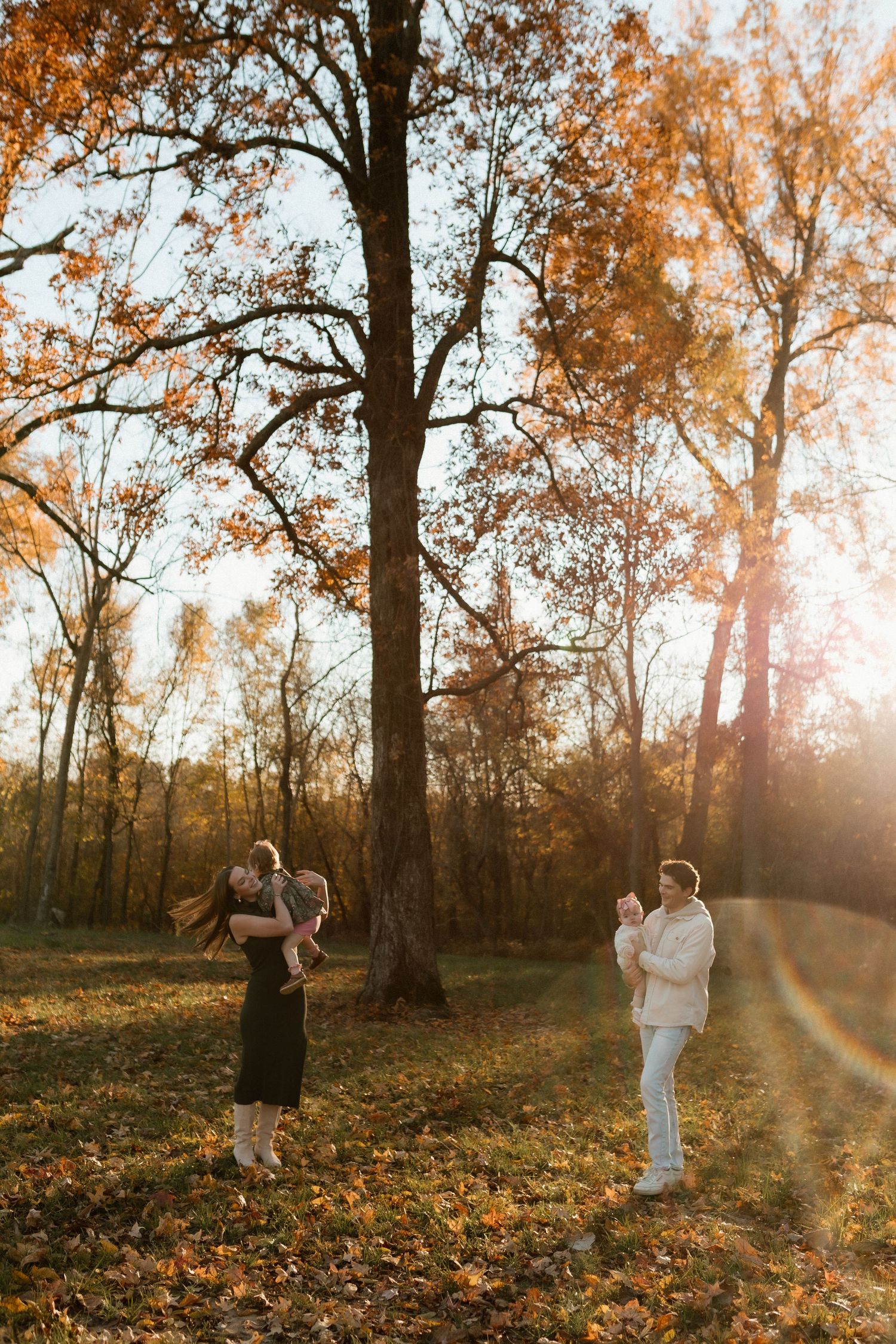 Family enjoying autumn sunset in a park, mother lifting child while father watches among colorful fall trees.