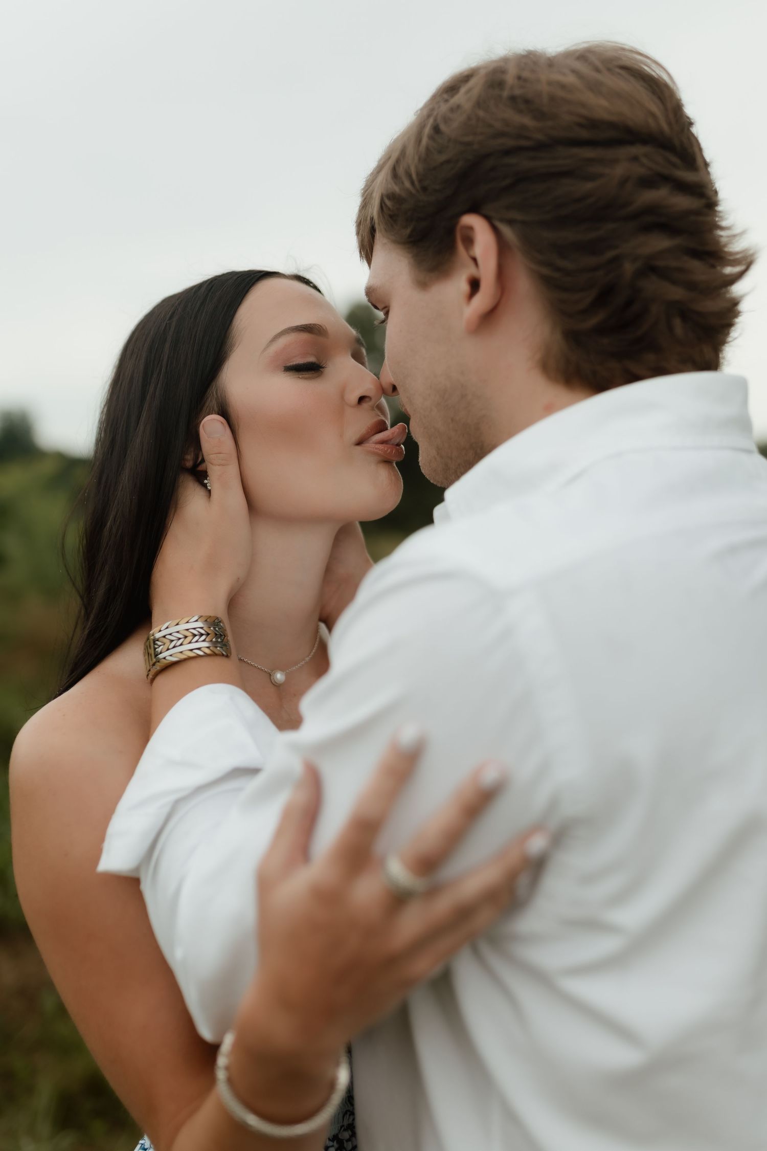 Couple sharing a passionate kiss outdoors, woman with dark hair wearing silver jewelry and white dress.