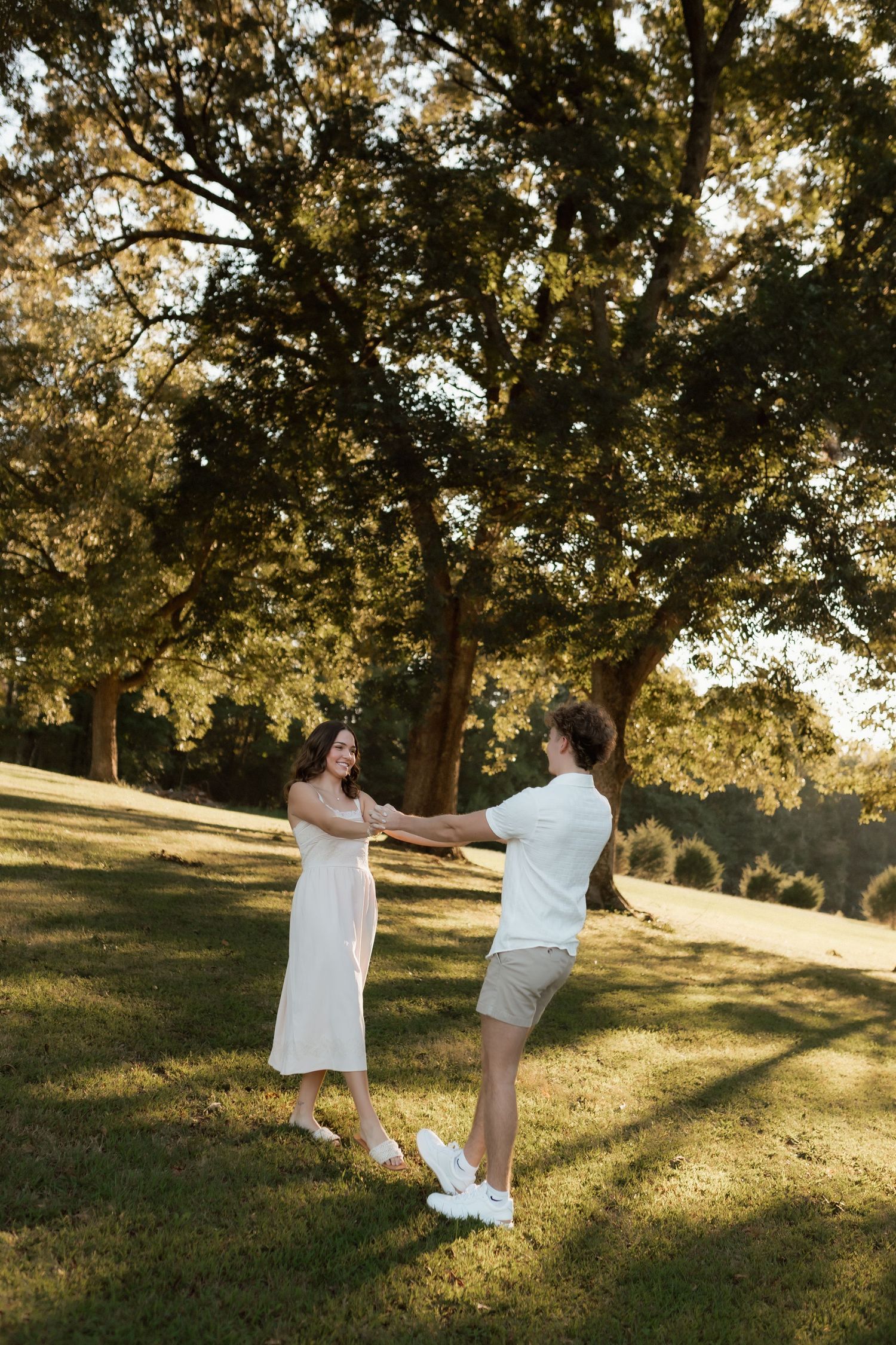 A couple holds hands and dances joyfully on a grassy lawn beneath a large oak tree in warm golden sunlight.
