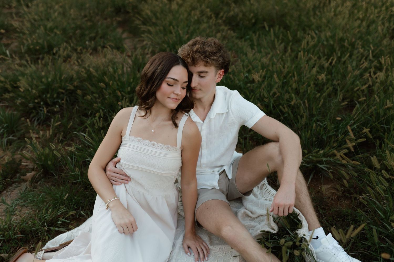 Couple sitting together in a grassy field at sunset, woman leaning into mans embrace outdoors.