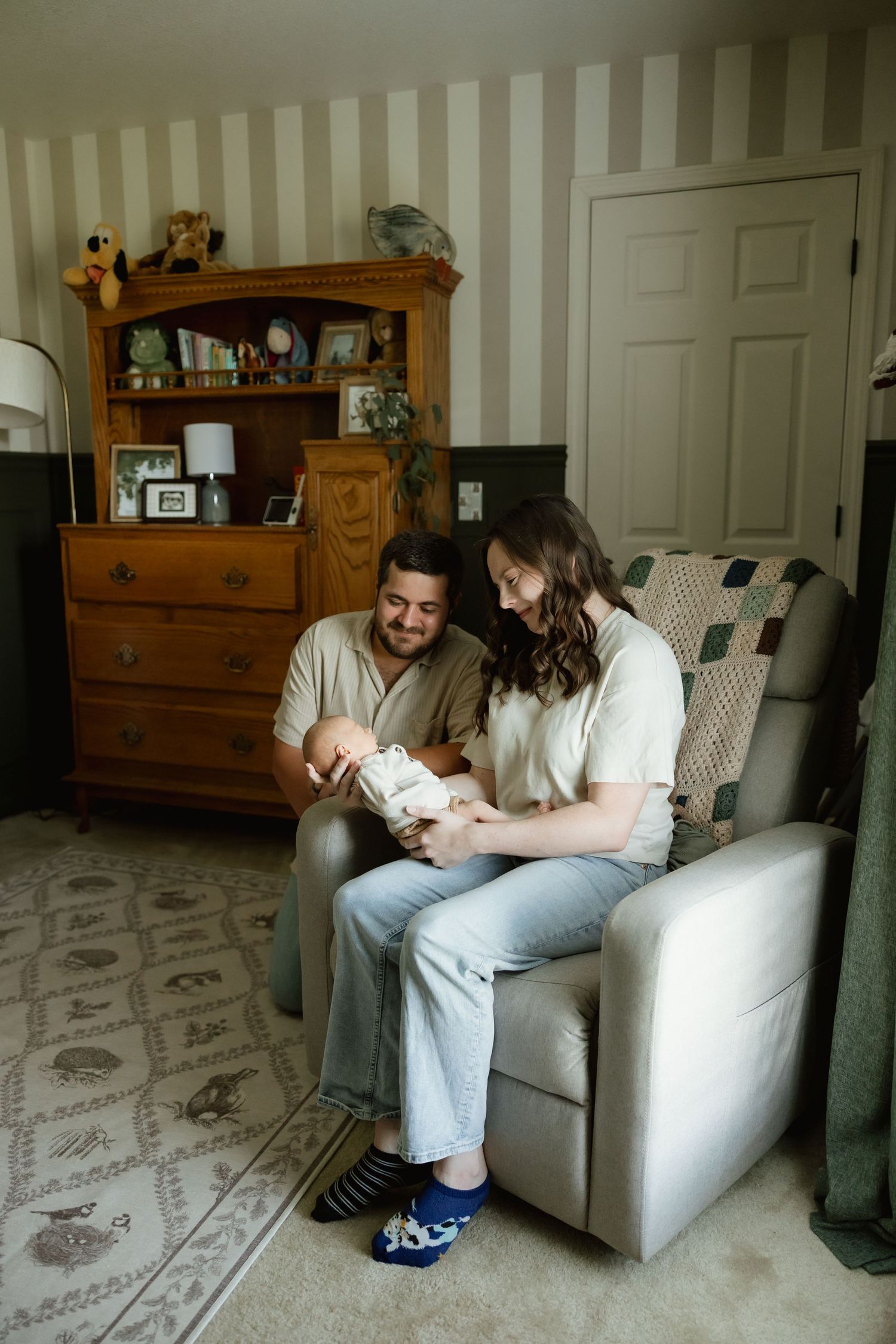 Happy couple admiring their newborn baby while seated in a gray recliner in a cozy nursery.