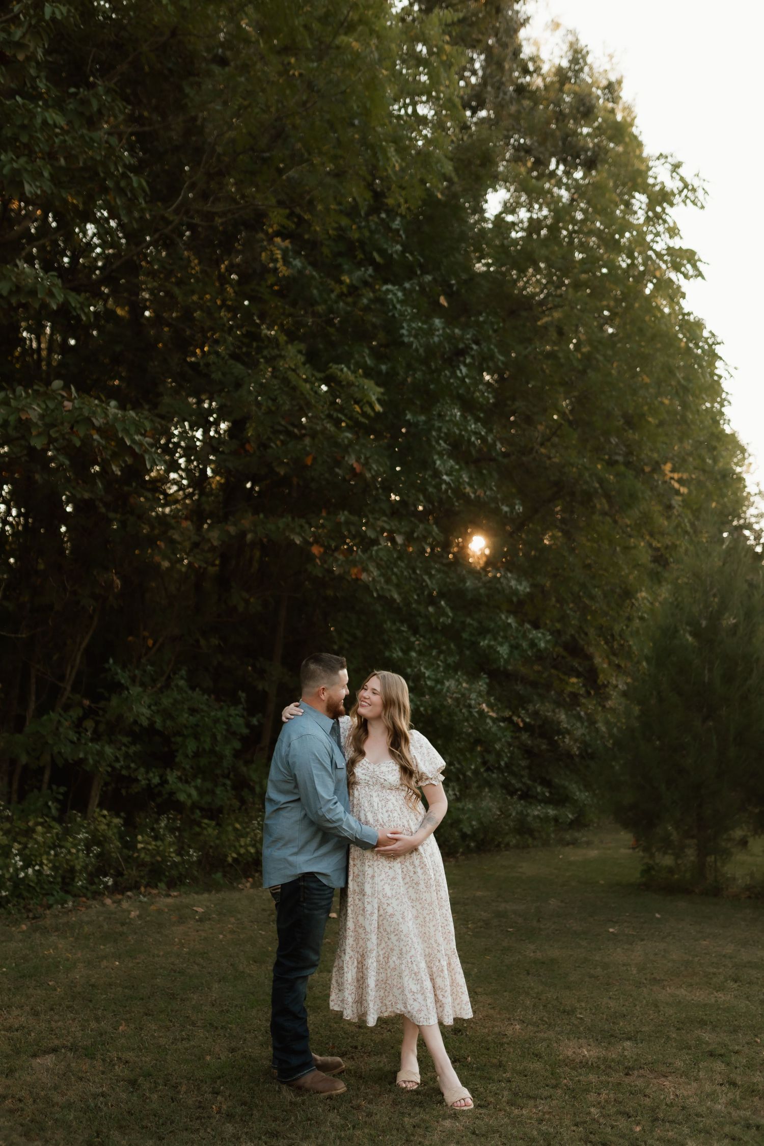 Couple sharing tender moment in garden at sunset, woman in floral dress cradling baby bump.