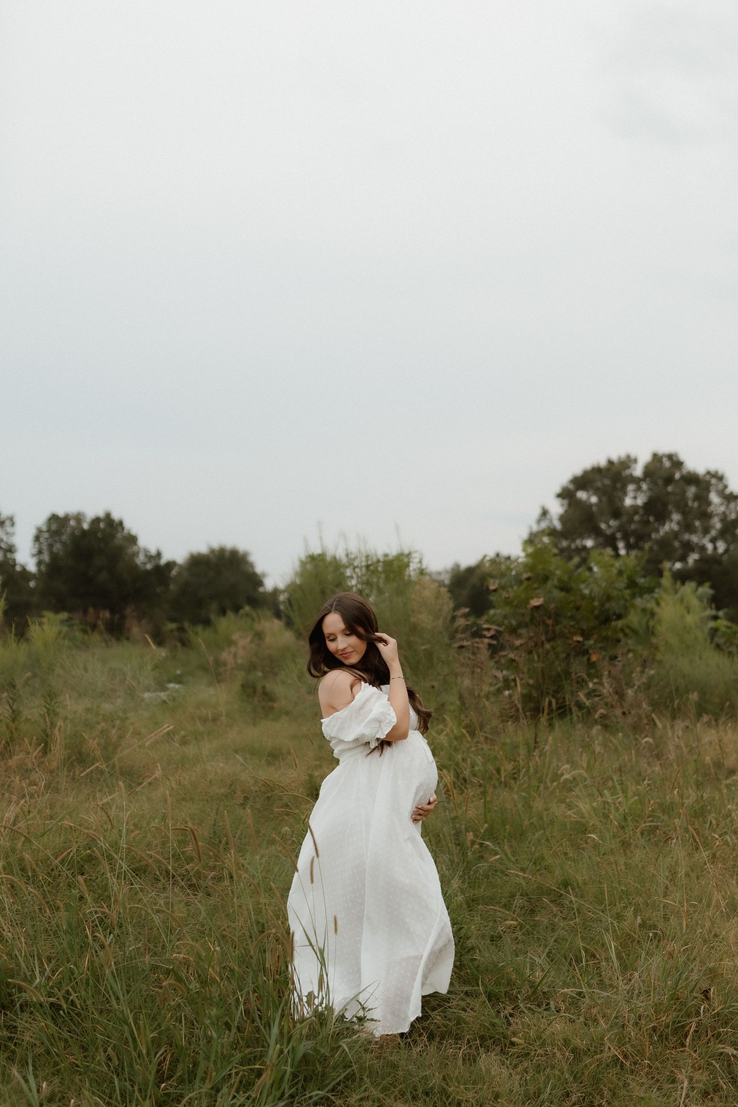 Pregnant woman in flowing white dress twirls gracefully in a lush green outdoor field.