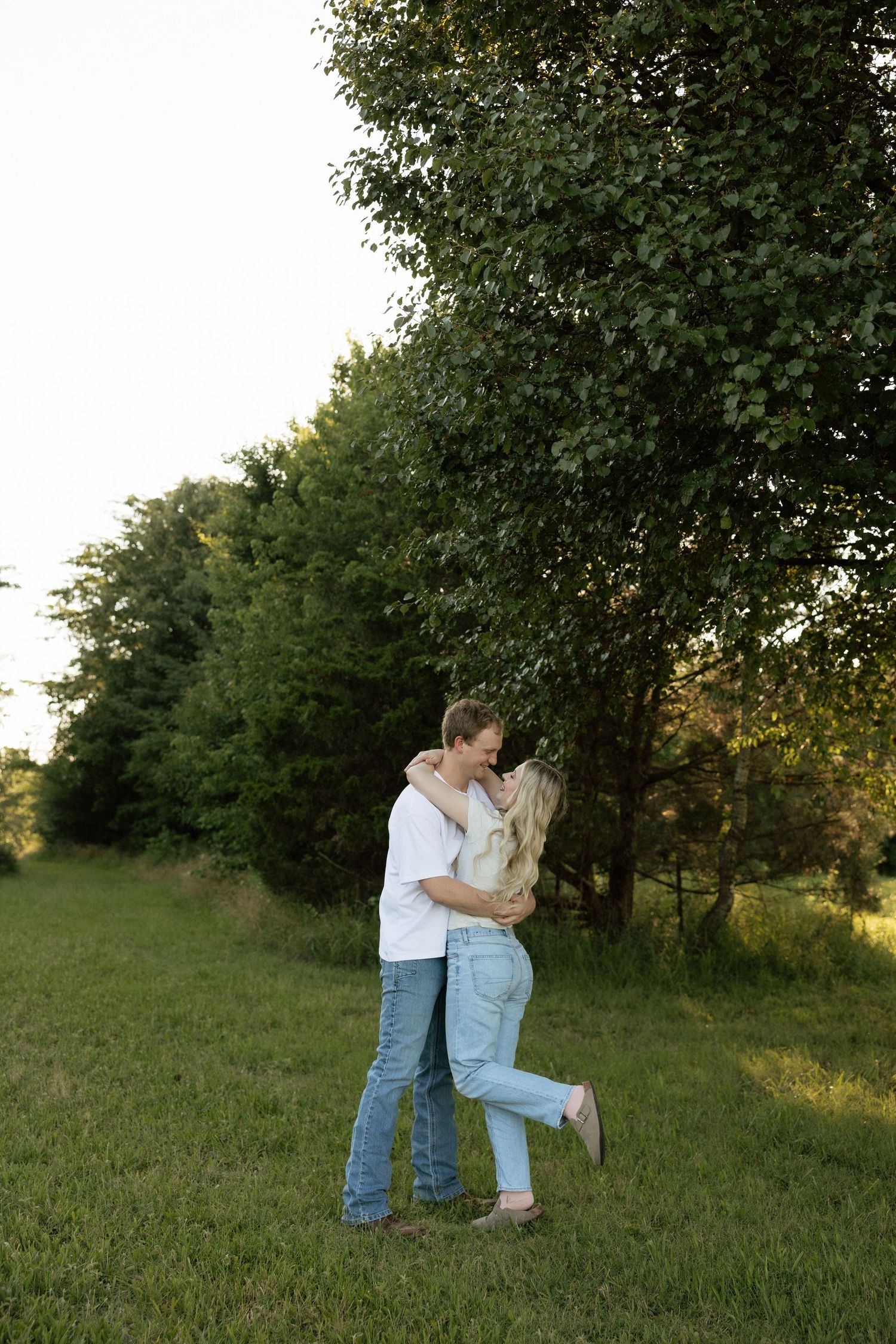 Couple sharing a romantic kiss while embracing in green field surrounded by trees during golden hour session.