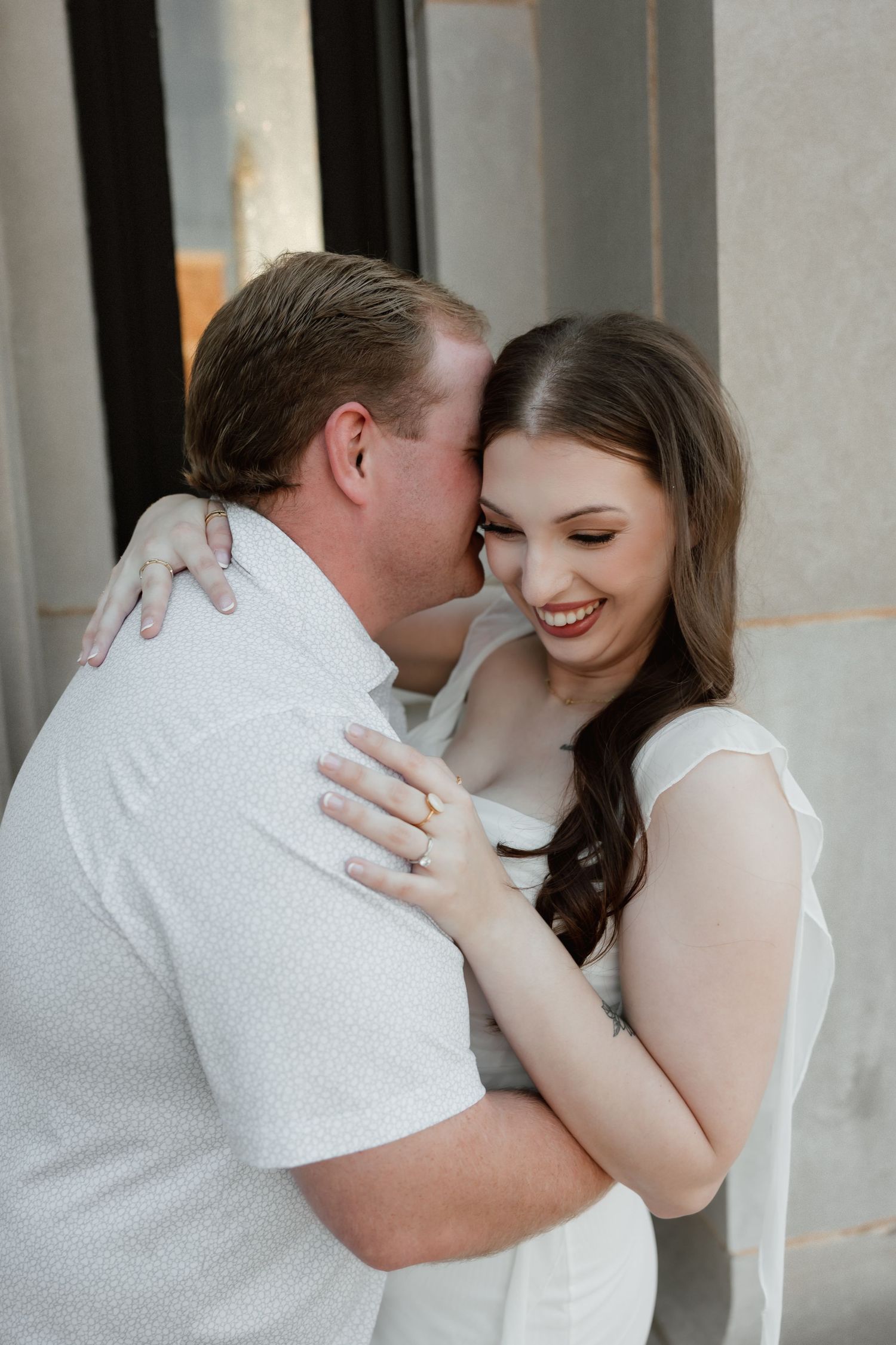 A joyful woman in white laughs as her partner embraces her during an engagement photo session.