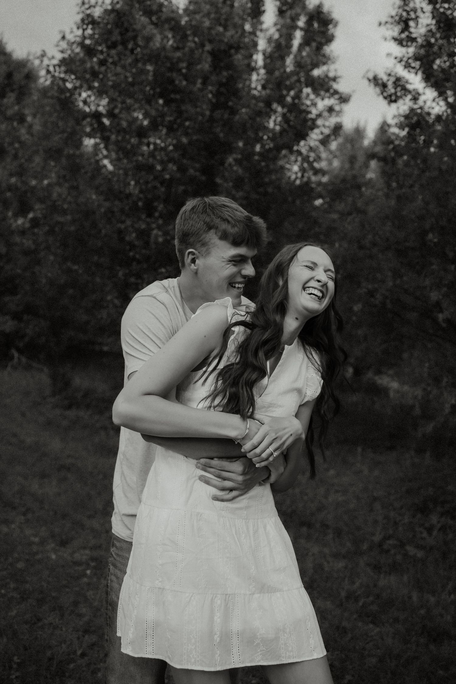 Black and white photo of a couple laughing joyfully as the man embraces the woman from behind outdoors.