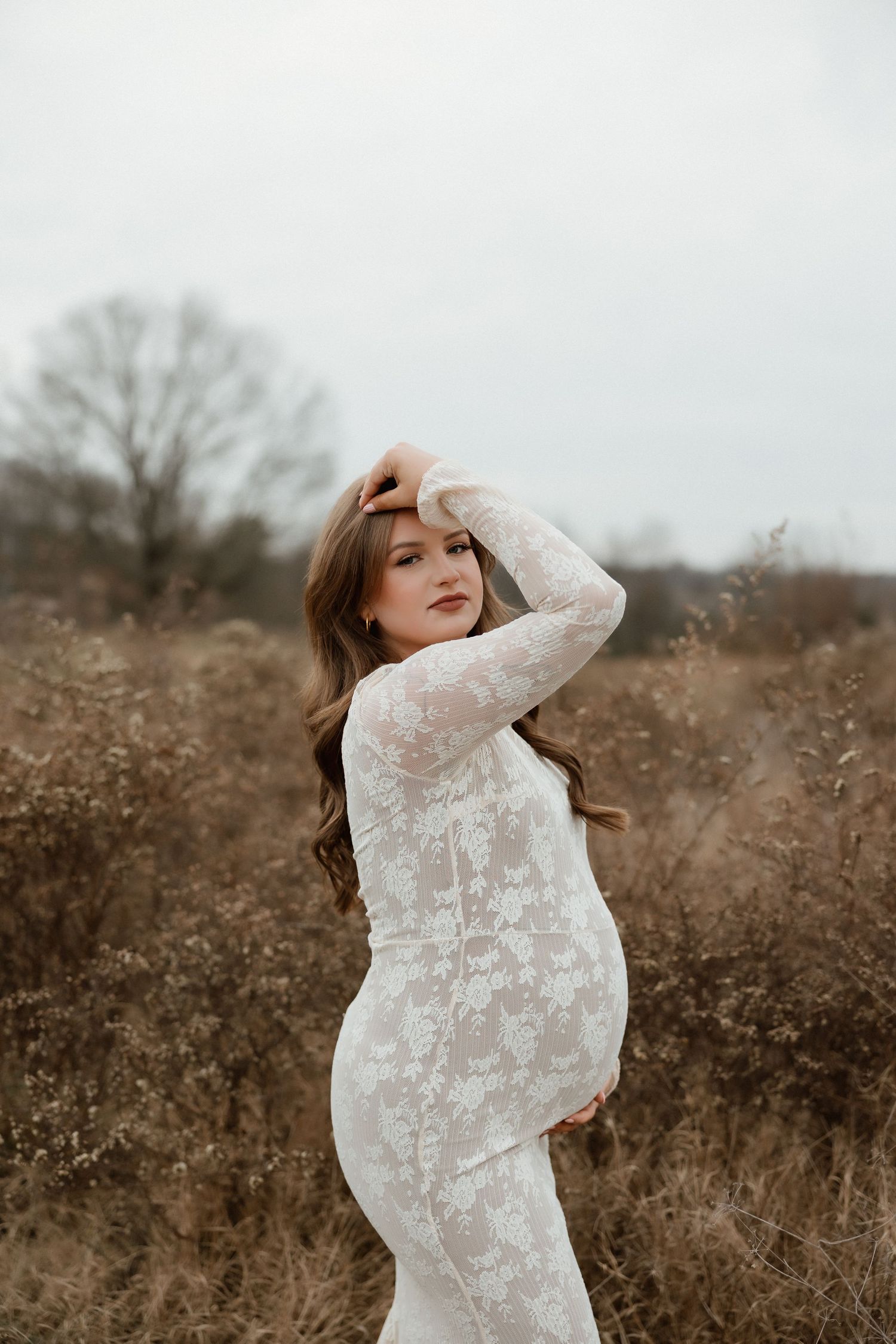 Pregnant woman in white lace maternity gown posing in golden field, hand in hair, looking over shoulder.