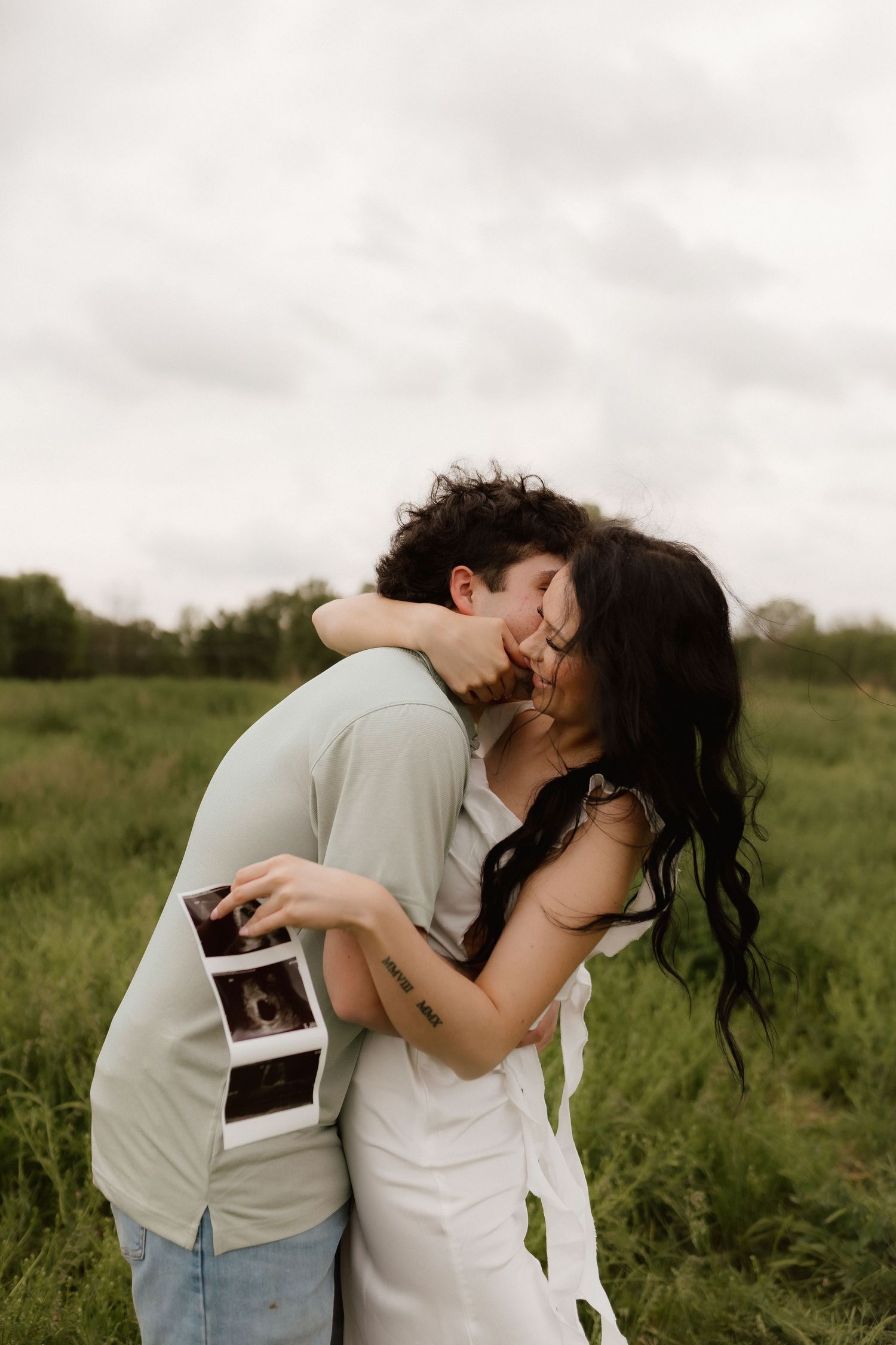 Couple sharing a kiss in green field during pregnancy announcement shoot, holding ultrasound photos between them.