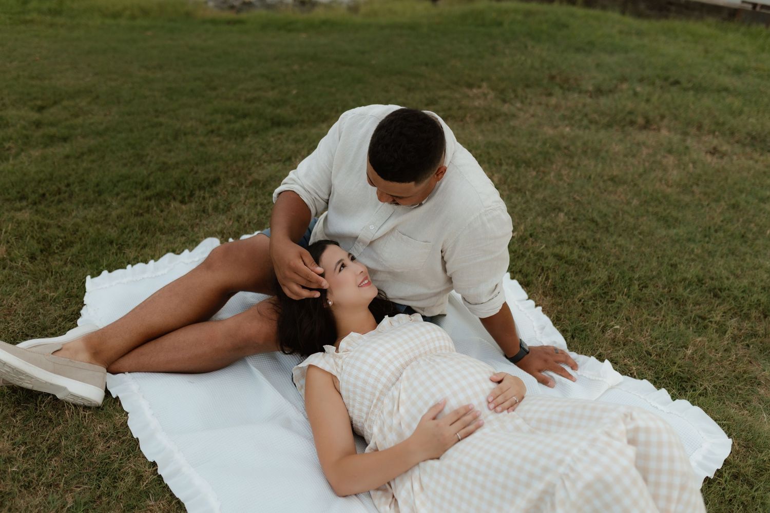 Pregnant couple relaxing on white blanket on grass, man tenderly looking at woman lying down.