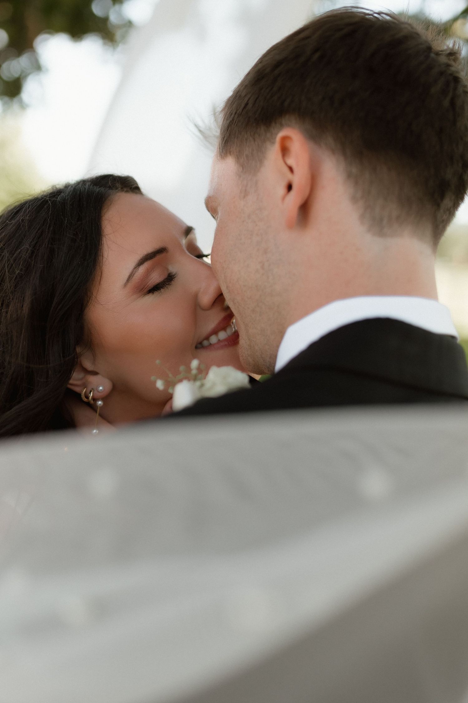 Close-up romantic moment of smiling bride leaning in to kiss groom outdoors with soft bokeh background.