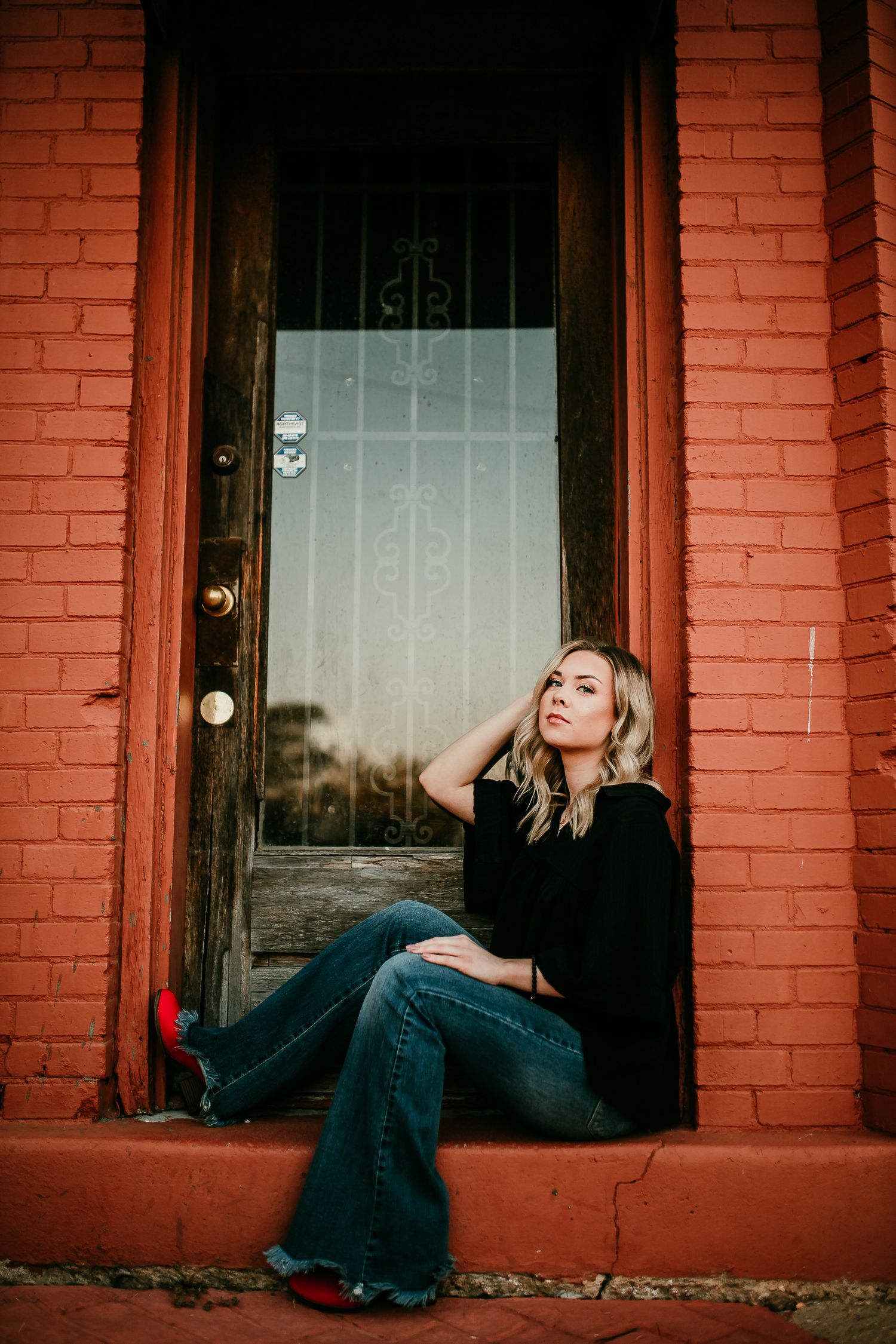 Blonde woman in black top and flared jeans sitting in vintage red brick doorway.