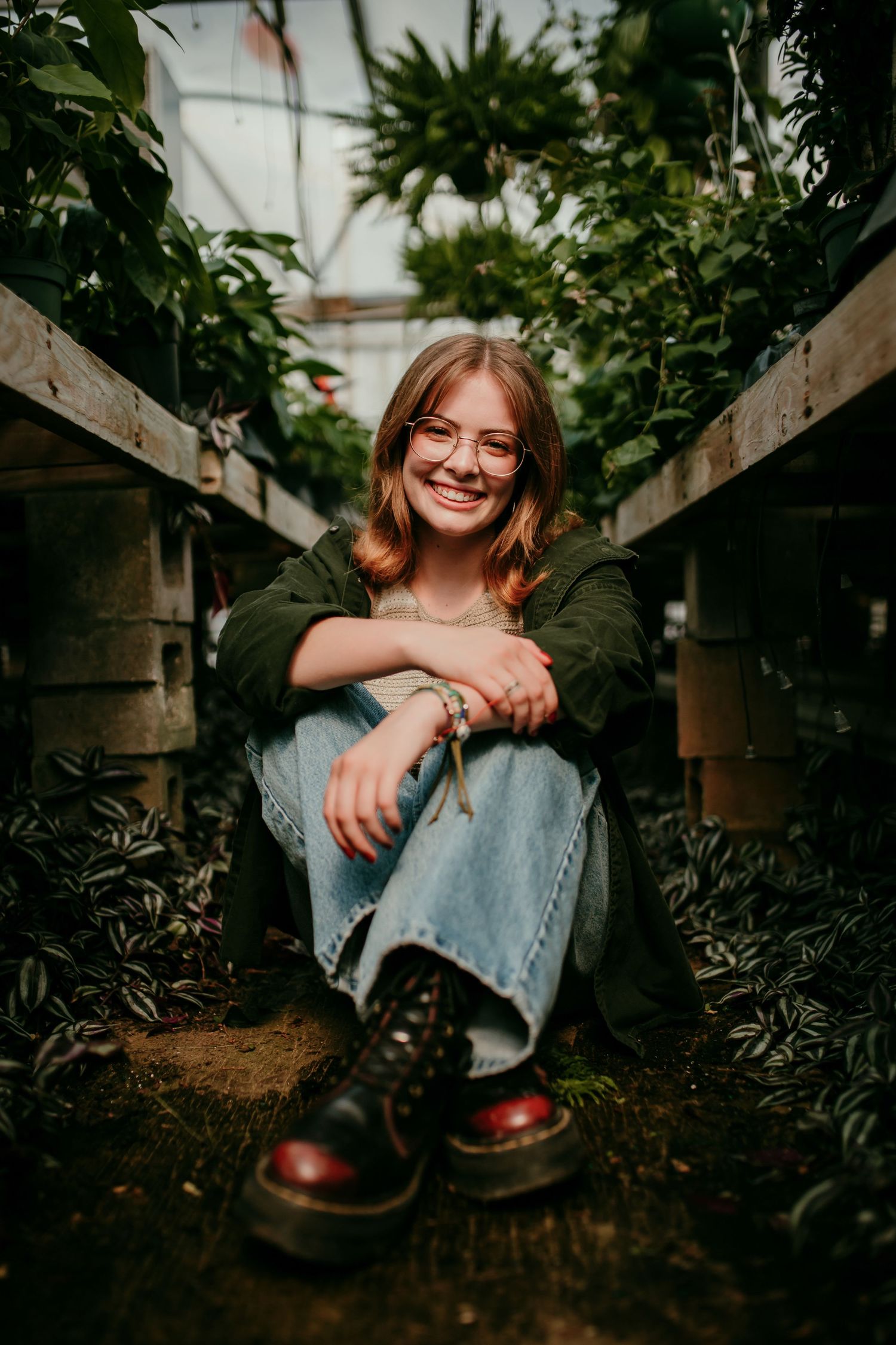 Smiling teenage girl with glasses and Dr. Martens boots sits cross-legged on greenhouse floor among plants.