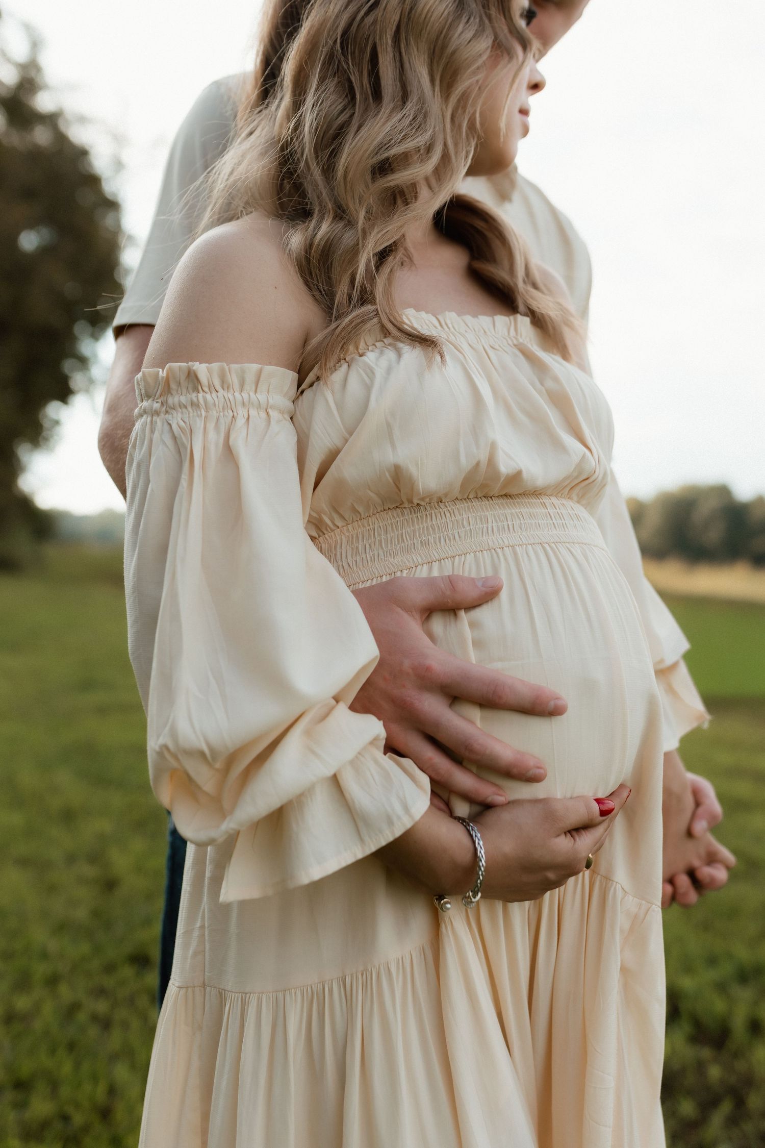Close-up of couple cradling pregnant belly in cream off-shoulder dress outdoors.