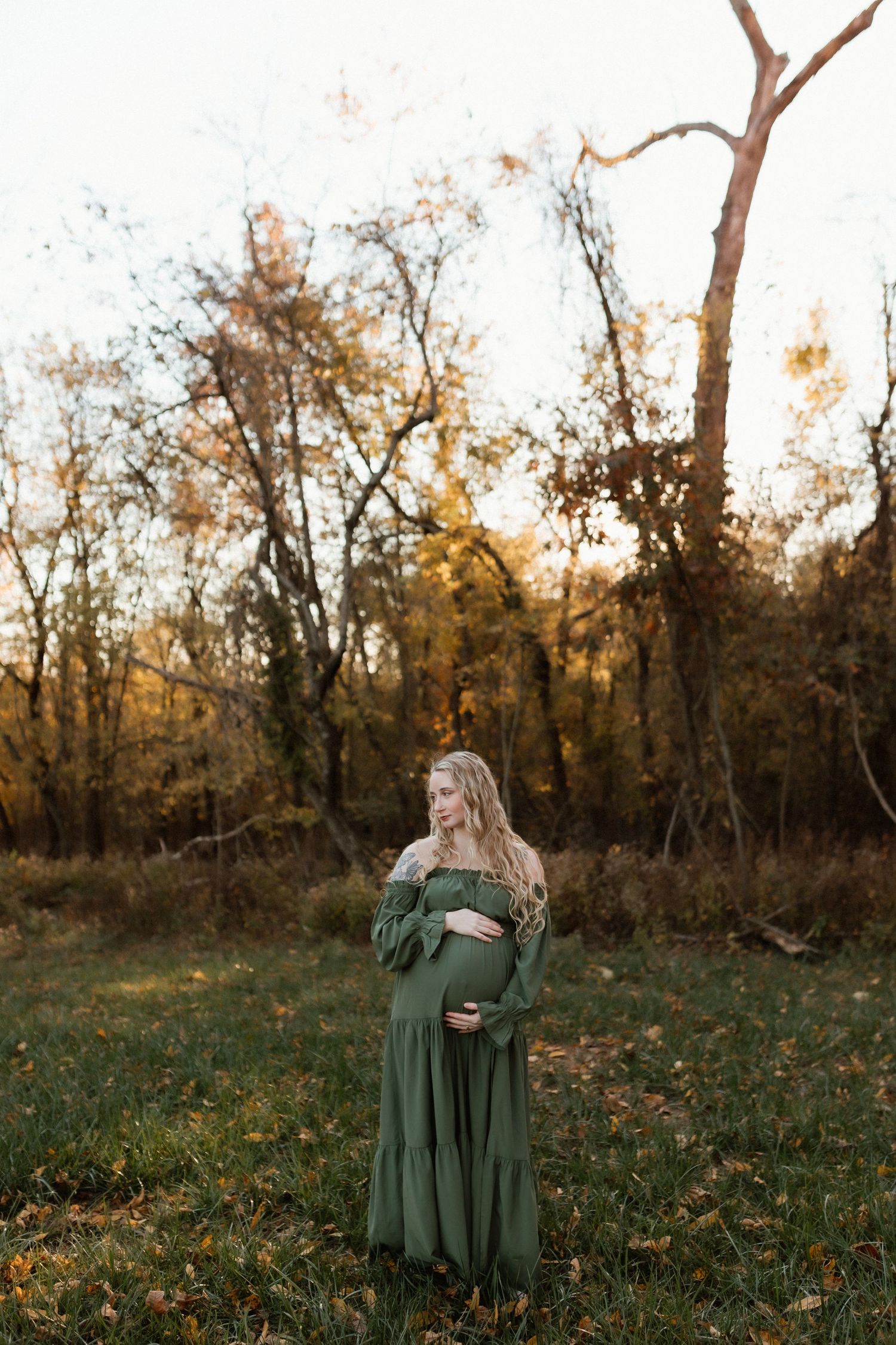 Pregnant woman in a flowing green off-shoulder gown standing in an autumn forest during golden hour.
