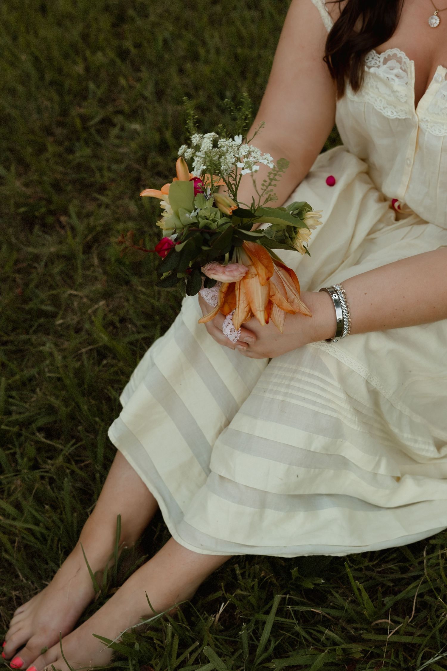 Barefoot woman in cream striped dress sitting on grass holding a small bouquet of flowers.